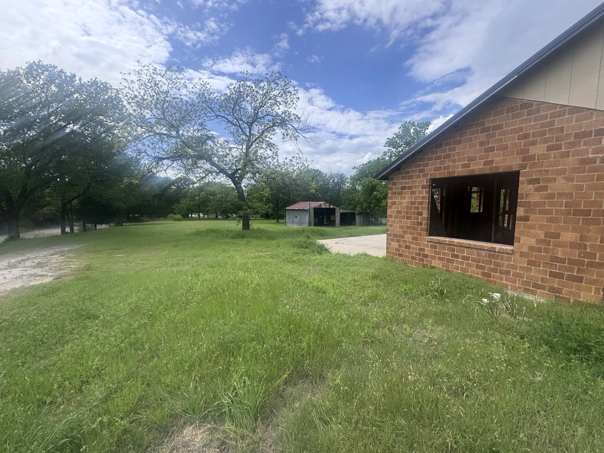 Brick exterior with a large open window, featuring a substantial grassy area, mature trees, and a concrete pad
