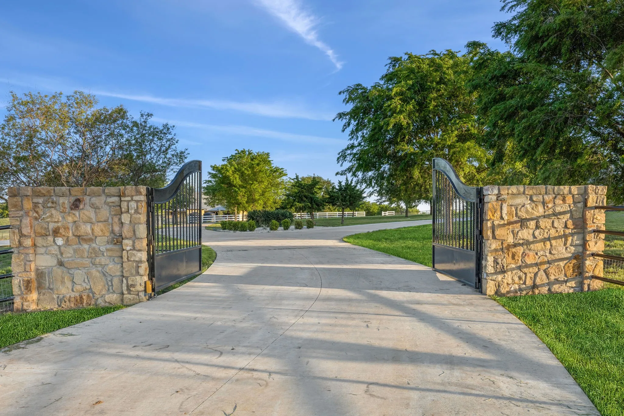 Stone pillar entryway featuring a black wrought iron gate, a concrete driveway, and surrounding green lawn with mature trees