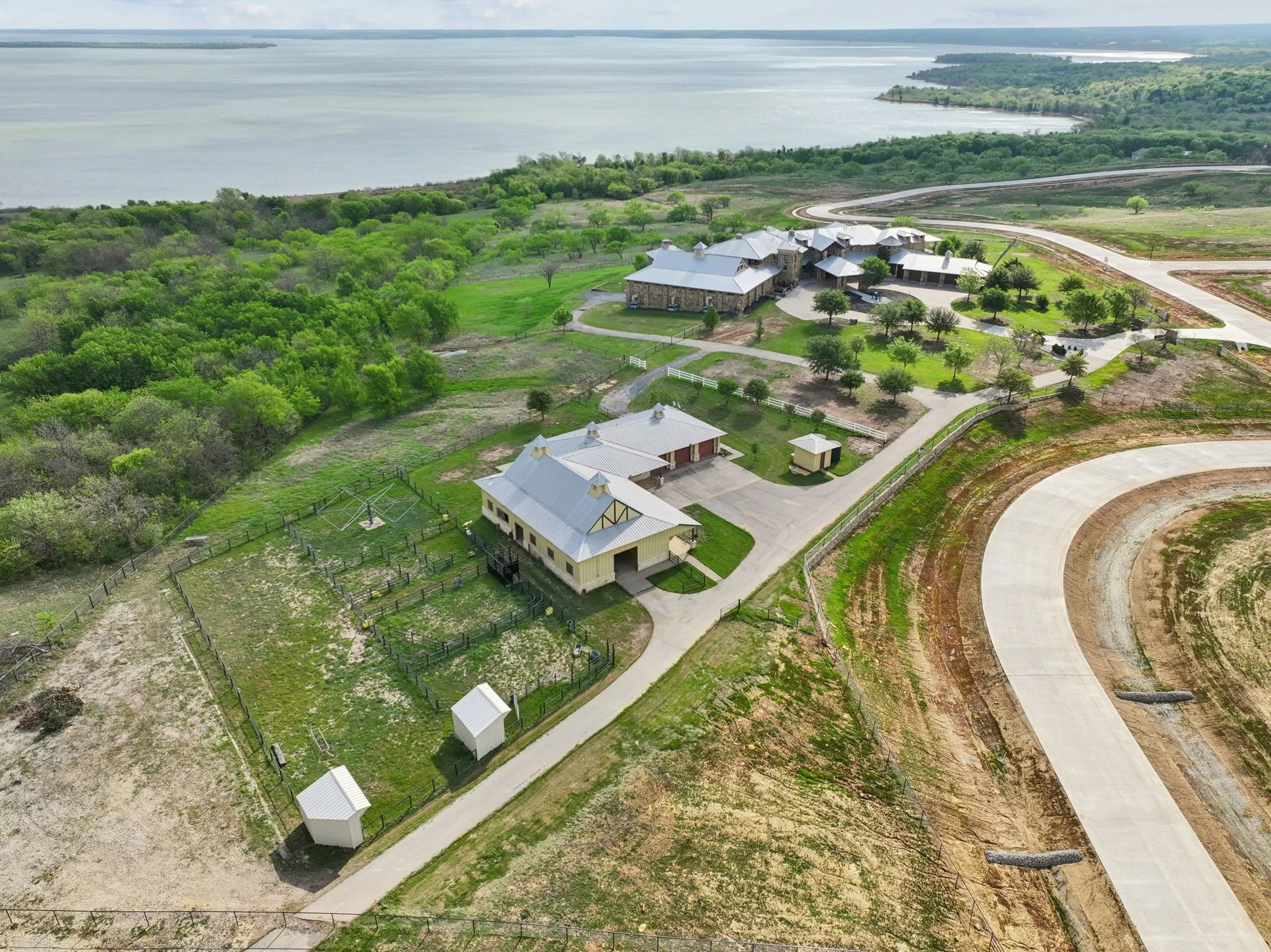 Expansive waterfront property featuring multiple structures, mature trees, concrete driveways, and a concrete perimeter road