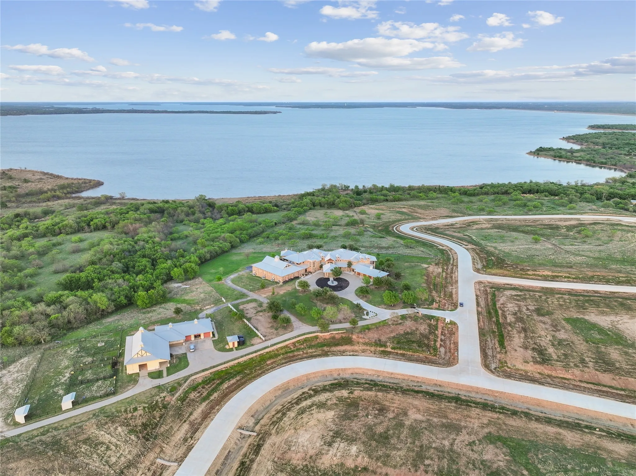 Expansive property featuring a large body of water, a main residence with a circular driveway, an additional structure with multiple garage bays, undeveloped land, and paved roadways