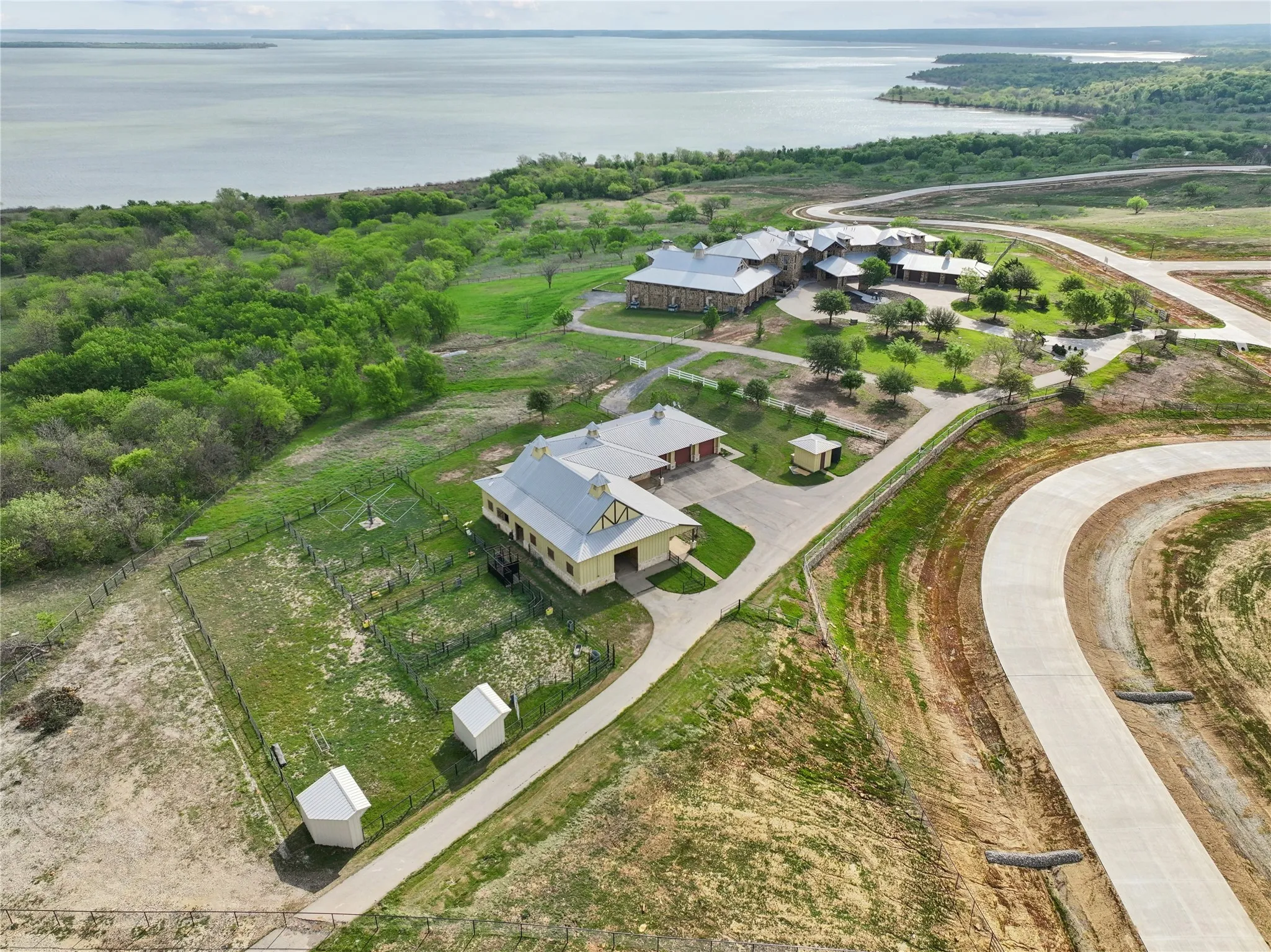 Expansive property featuring multiple structures, paved driveways, established landscaping, and a fenced area, bordered by a large body of water