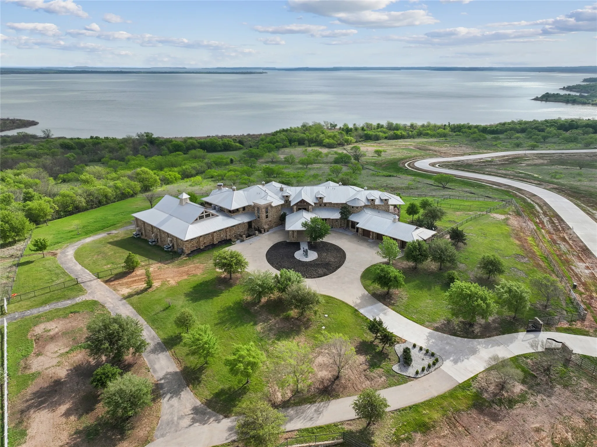 Expansive stone estate featuring a circular drive, multiple outbuildings, and extensive green space, all bordered by a large body of water.
