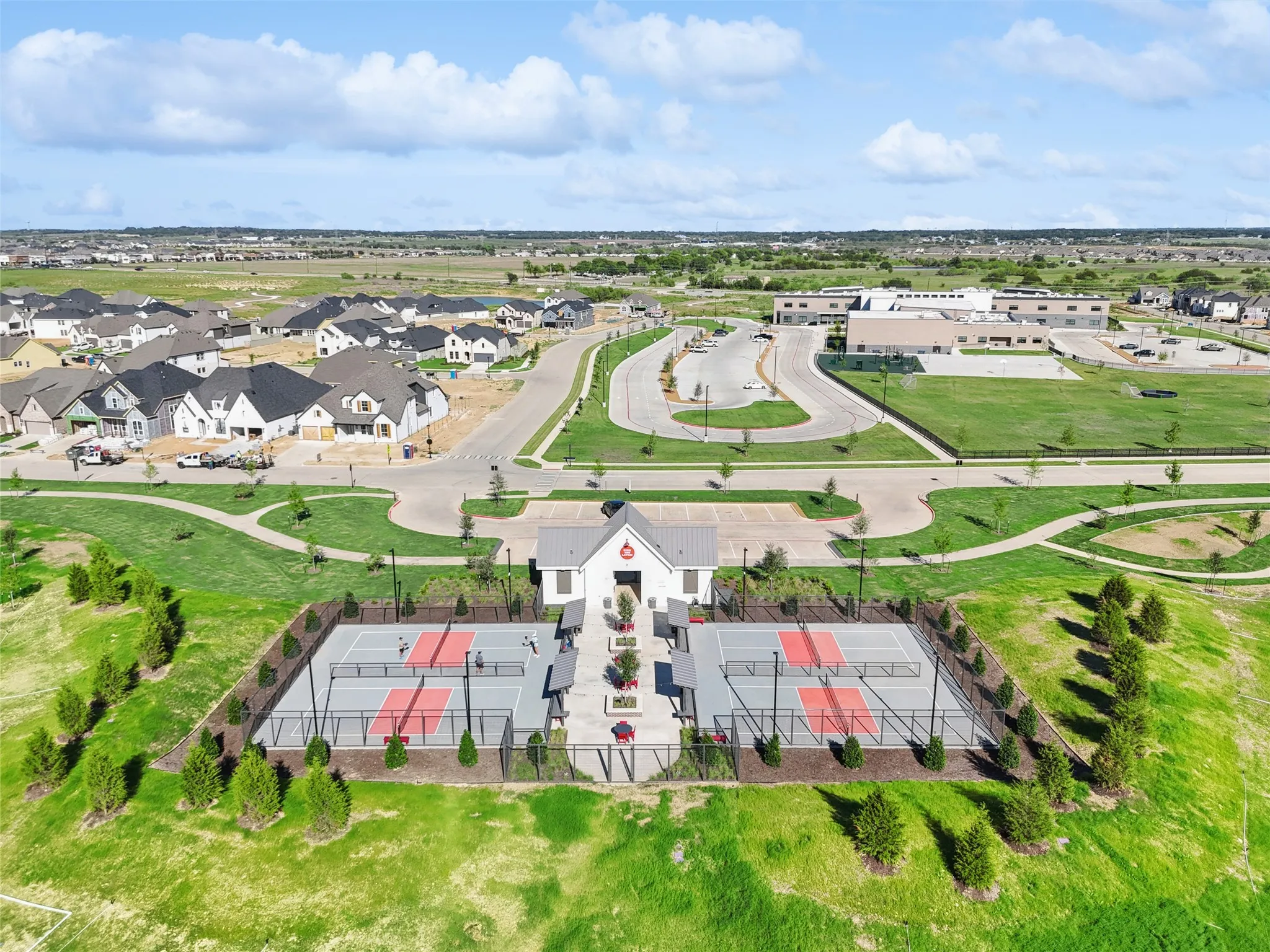 Community recreational facility featuring a white structure, two fenced sports courts with red and gray surfaces, surrounding landscaped grounds, and a paved pathway system