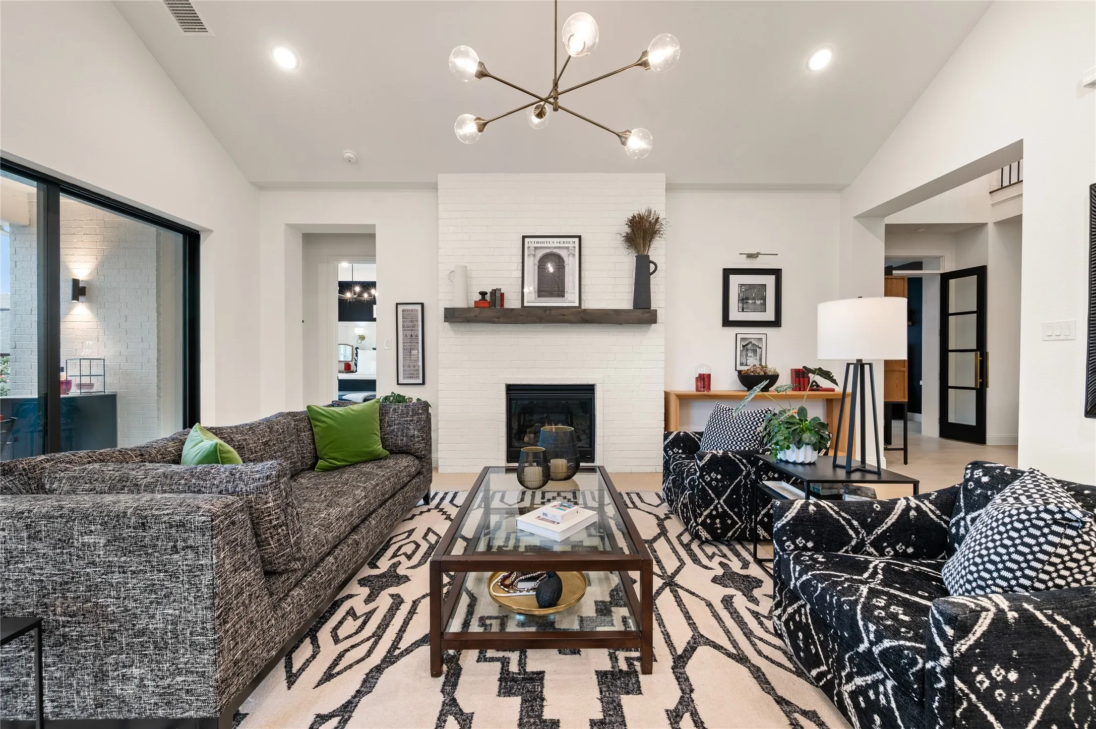 Spacious living area featuring expansive sliding glass doors overlooking a serene backyar, vaulted ceilings, light wood flooring, custom hardware and fixtures with a white painted brick fireplace with a dark wood mantel for contrast.