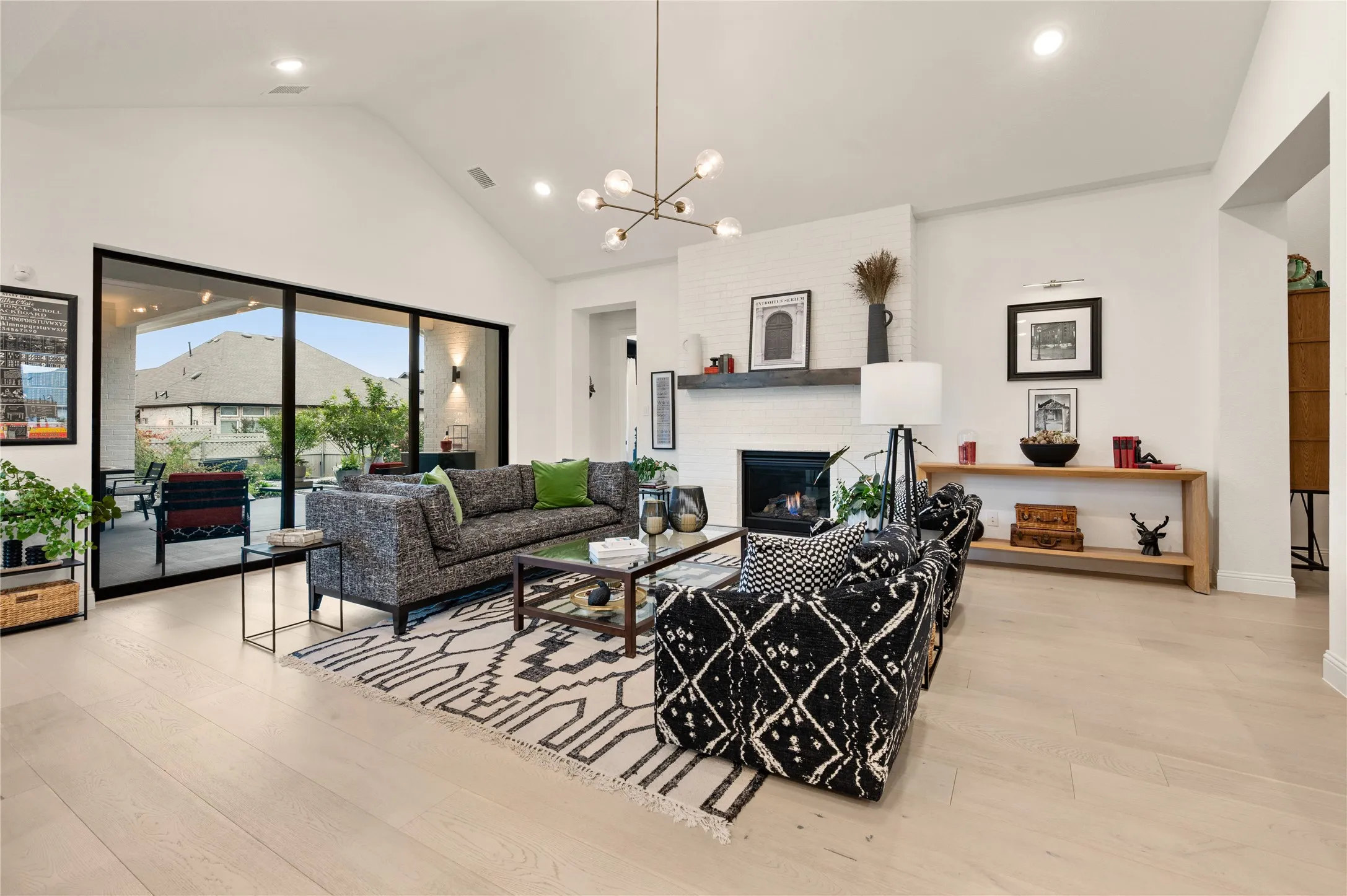 Spacious living area featuring expansive sliding glass doors overlooking a serene backyar, vaulted ceilings, light wood flooring, custom hardware and fixtures with a white painted brick fireplace with a dark wood mantel for contrast.