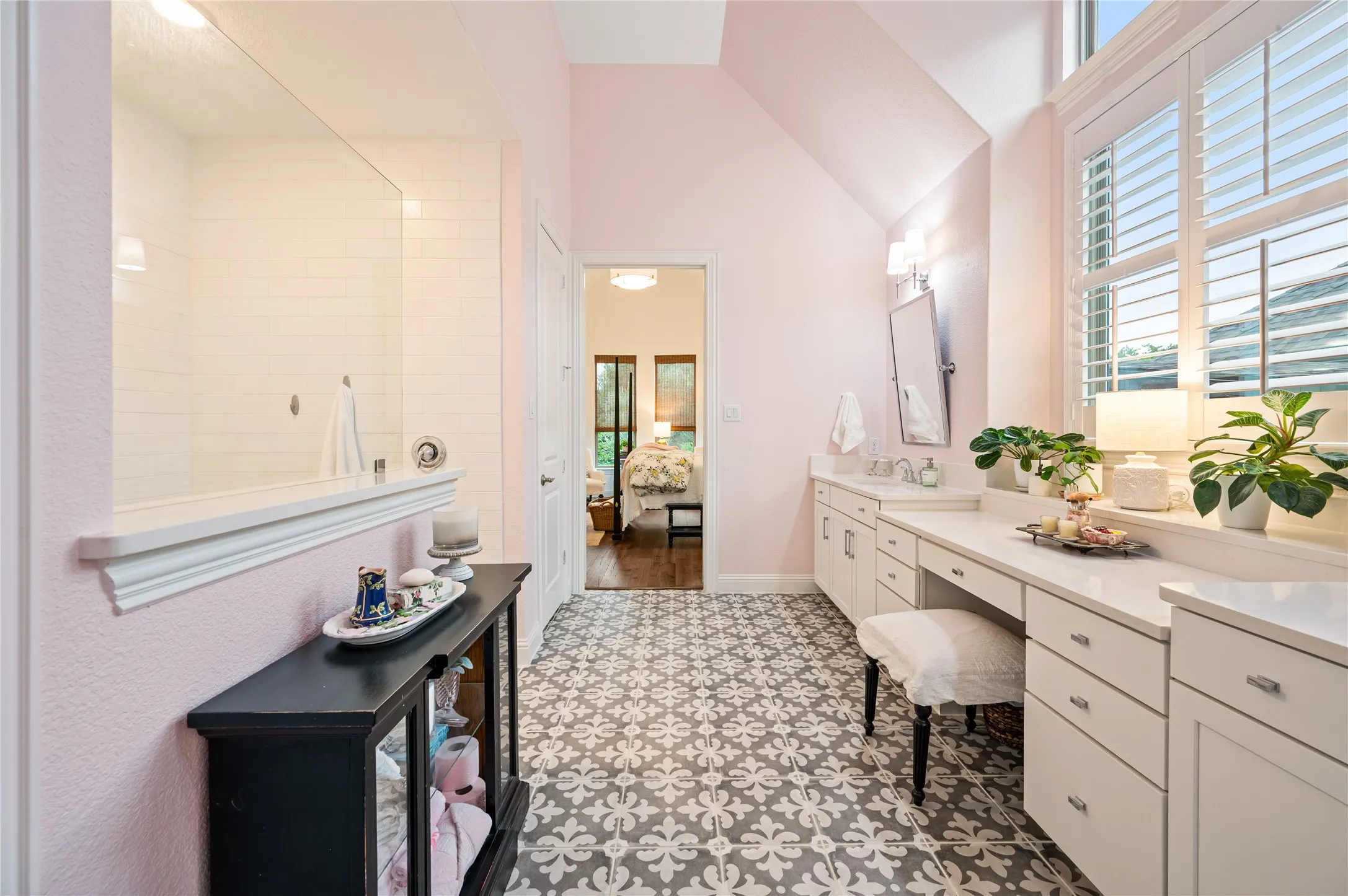Spacious bathroom featuring twin vanities with white cabinetry and quartz countertops, patterned floor tiles, and a large ADA accessible shower with a glass enclosure and walk in custom closet leading to laundry