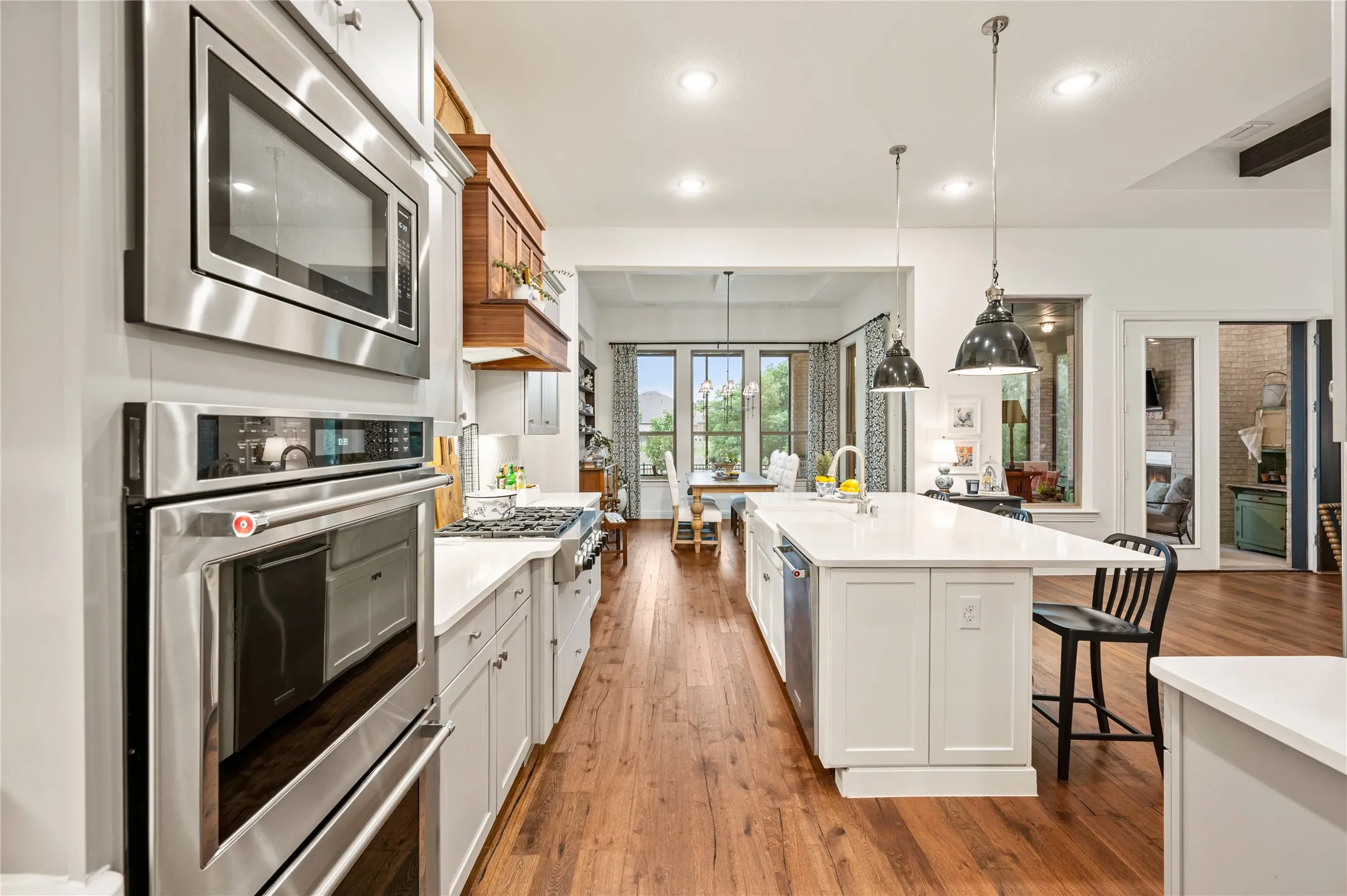 Gourmet kitchen with wood-finish flooring, stainless steel built-in double oven and microwave, white perimeter cabinetry, a gas range, and a large central island with pendant lighting and bar seating