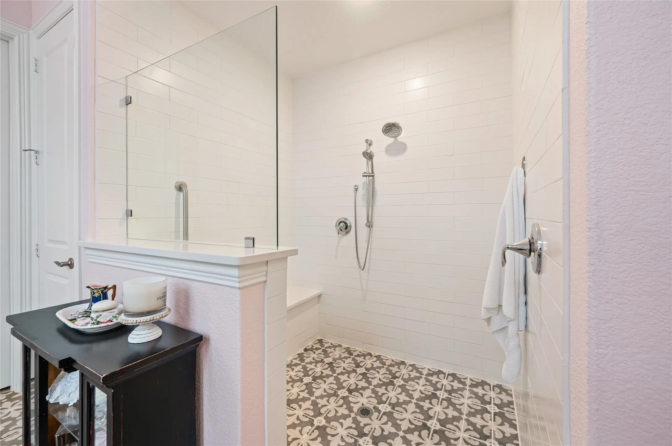 Spacious bathroom featuring twin vanities with white cabinetry and quartz countertops, patterned floor tiles, and a large ADA accessible shower with a glass enclosure and walk in custom closet leading to laundry