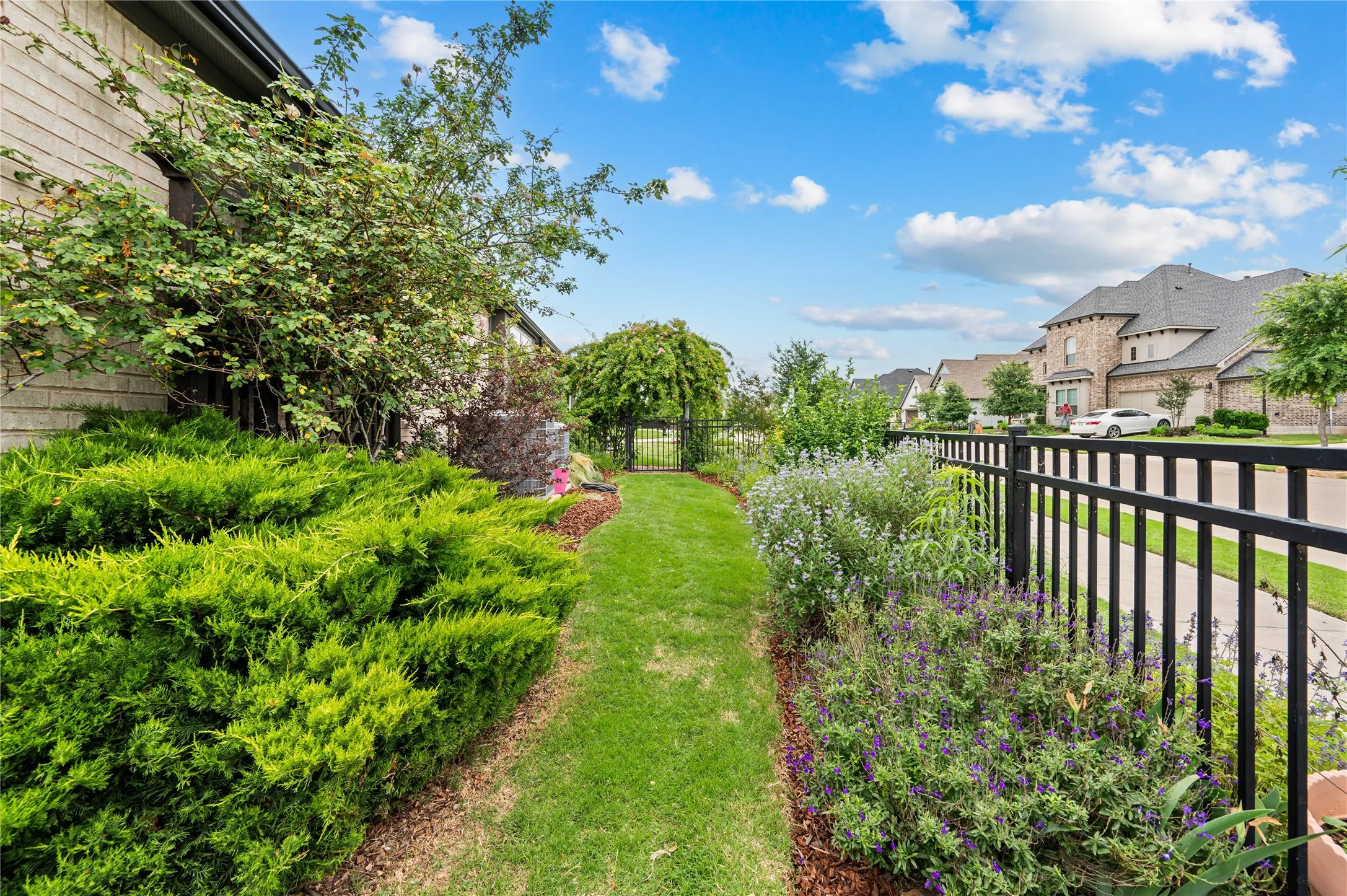 Manicured side yard featuring a lush lawn path, mature shrubbery, decorative garden beds, and a black wrought iron fence