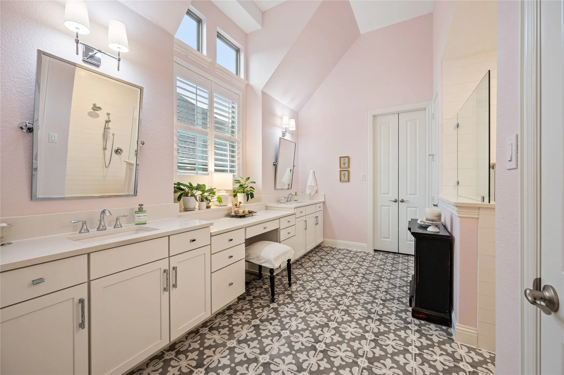 Spacious bathroom featuring twin vanities with white cabinetry and quartz countertops, patterned floor tiles, and a large ADA accessible shower with a glass enclosure and walk in custom closet leading to laundry