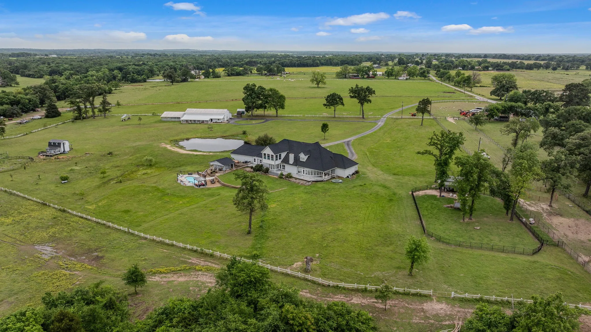 Expansive property featuring a white residence with a dark roof, a swimming pool, a pond, and multiple outbuildings