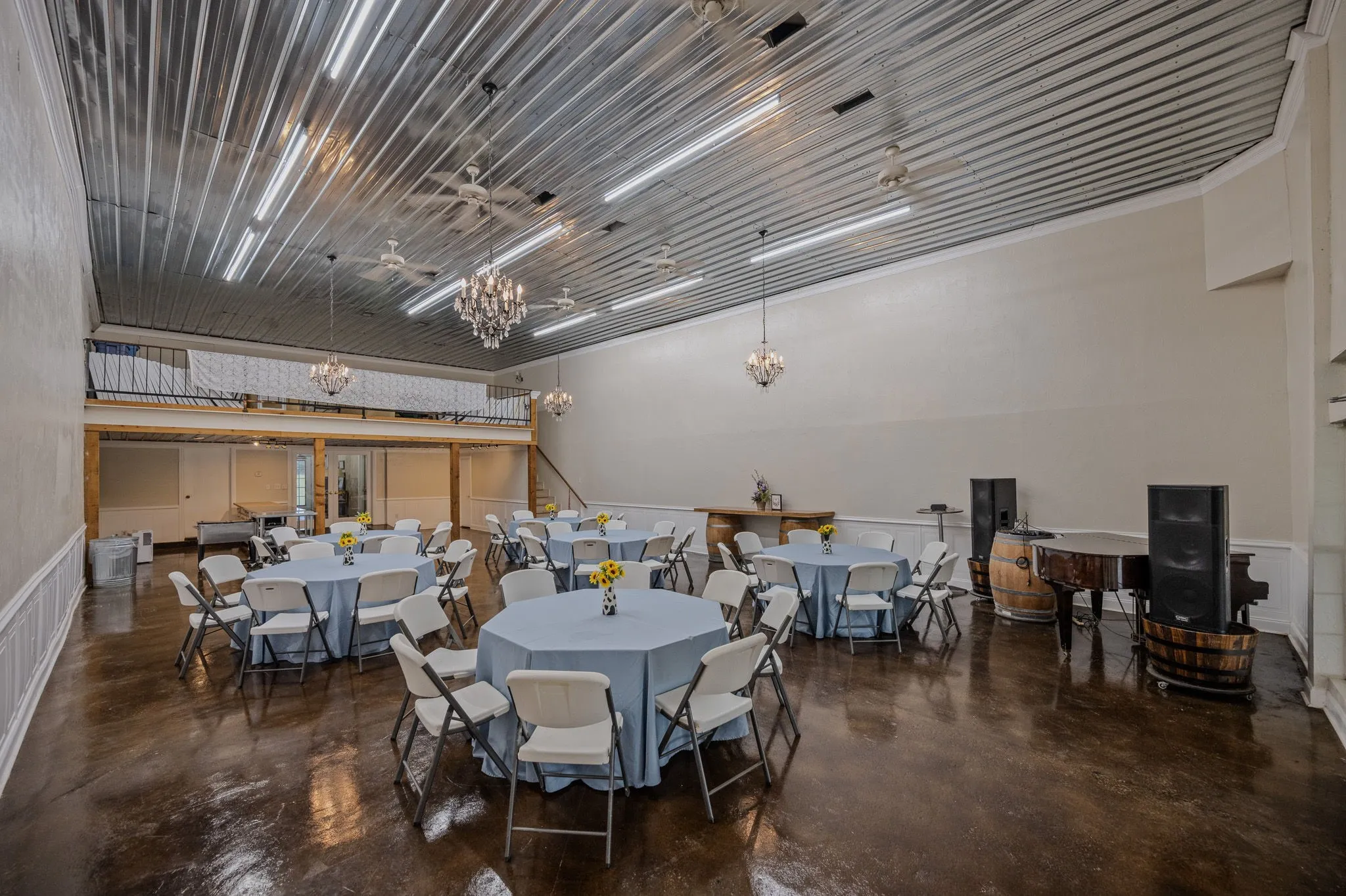 Expansive event hall featuring a high corrugated metal ceiling, polished concrete flooring, and a visible mezzanine