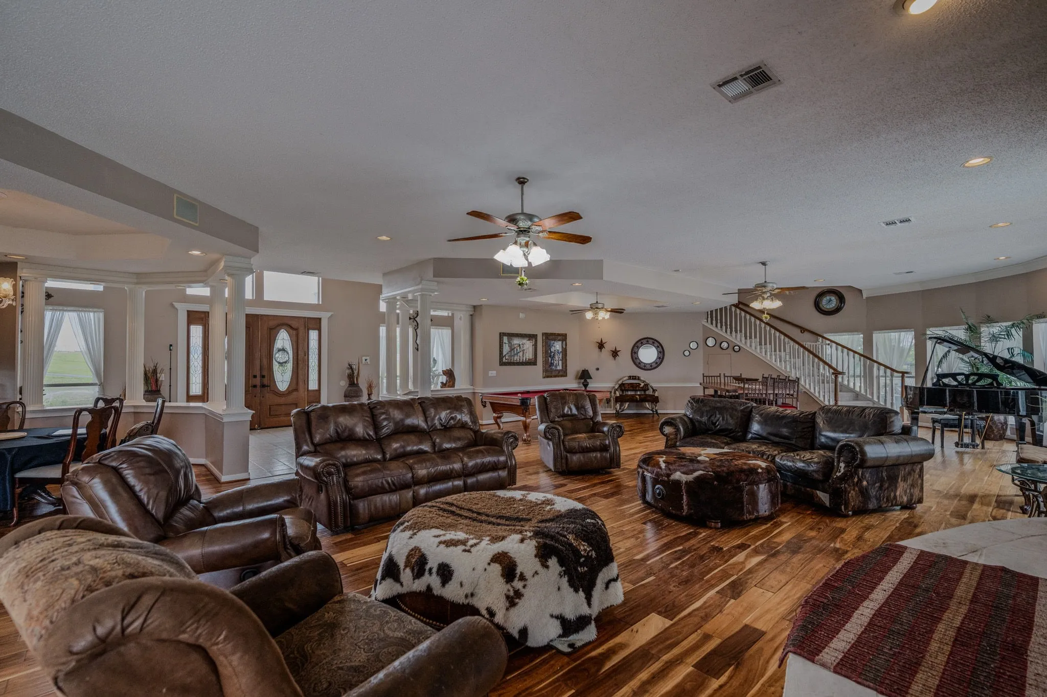 Expansive open-concept living area featuring rich wood-finish flooring, a grand staircase with white risers and wood treads, decorative columns, a front entry door with leaded glass, and multiple large windows