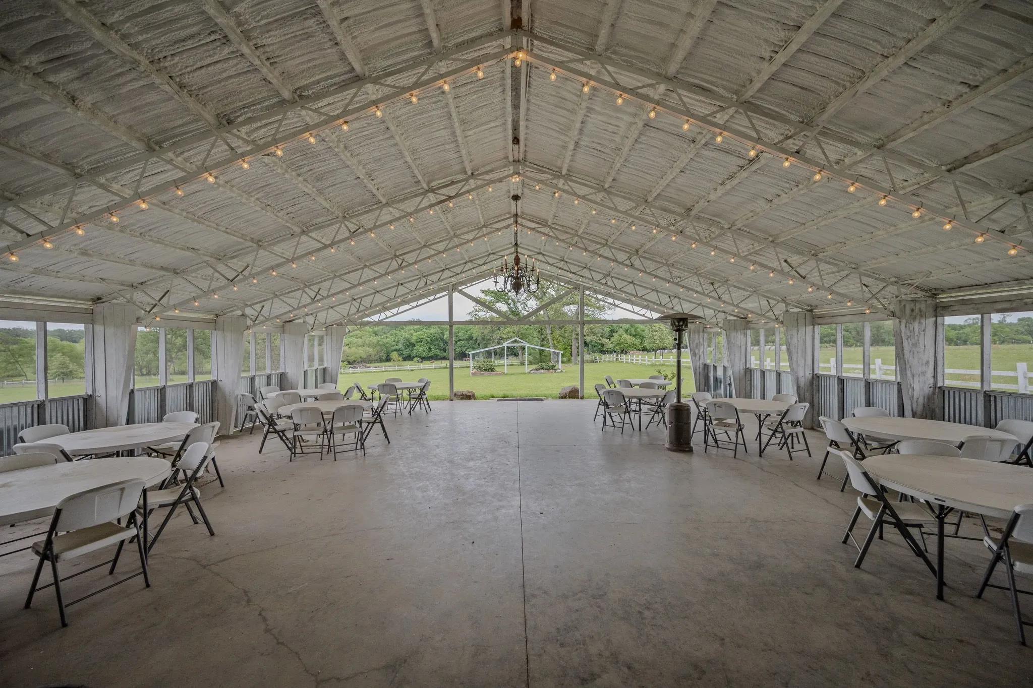 Spacious covered outdoor structure featuring a high vaulted ceiling with exposed metal trusses, string lighting, and a central chandelier