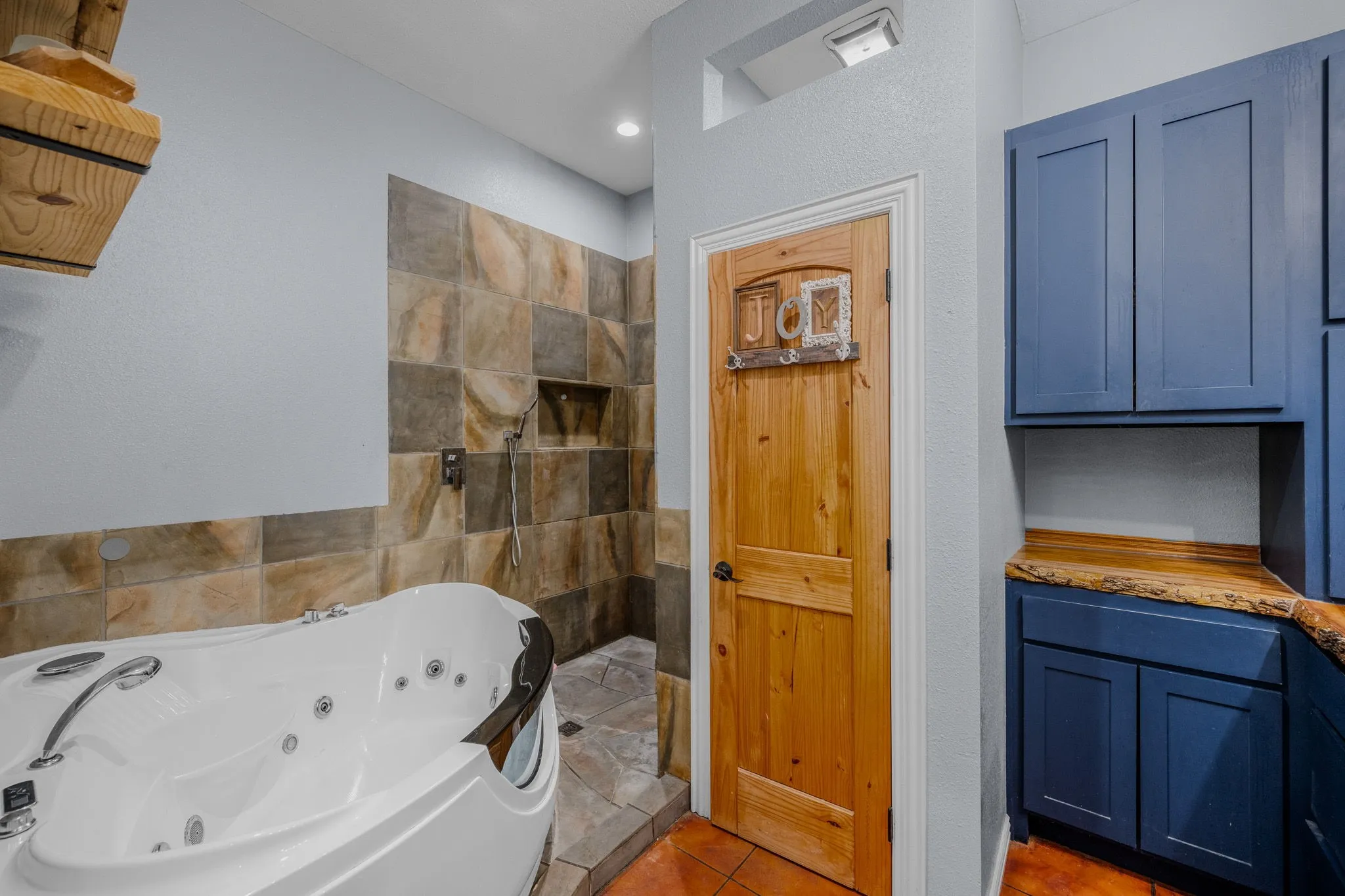 Luxurious soaking tub with chrome fixtures, a tiled shower enclosure with a recessed shelf, rustic wood-finish door, and custom blue cabinetry with a live-edge wood countertop