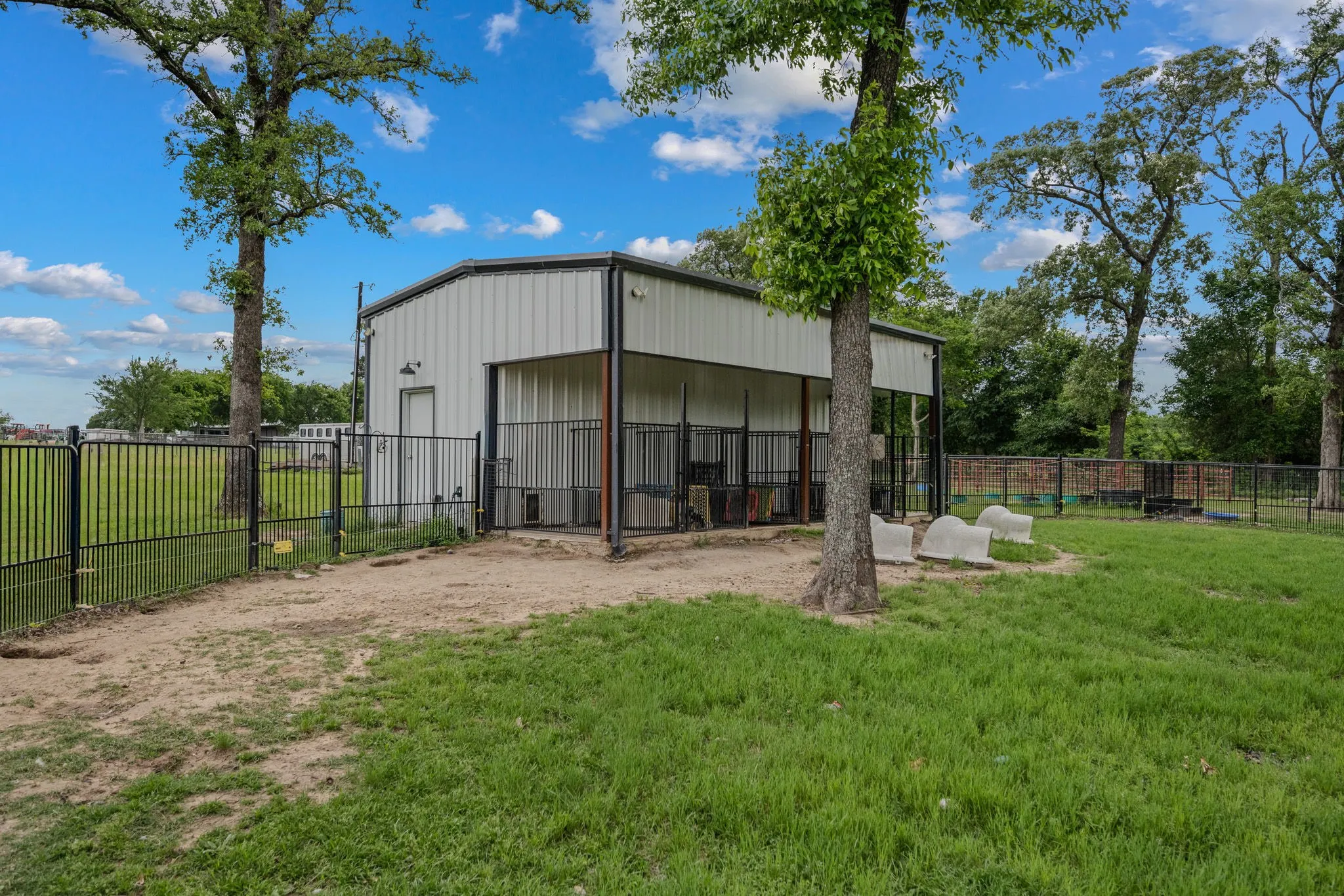 Metal building with covered outdoor stalls, black fencing, mature trees, and a spacious grassy area