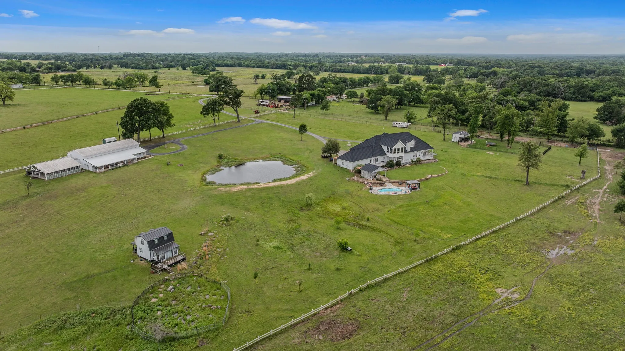 Expansive acreage featuring a main residence with a dark roof and light exterior, a swimming pool, a pond, an extended white outbuilding, and a separate small structure with a dark roof