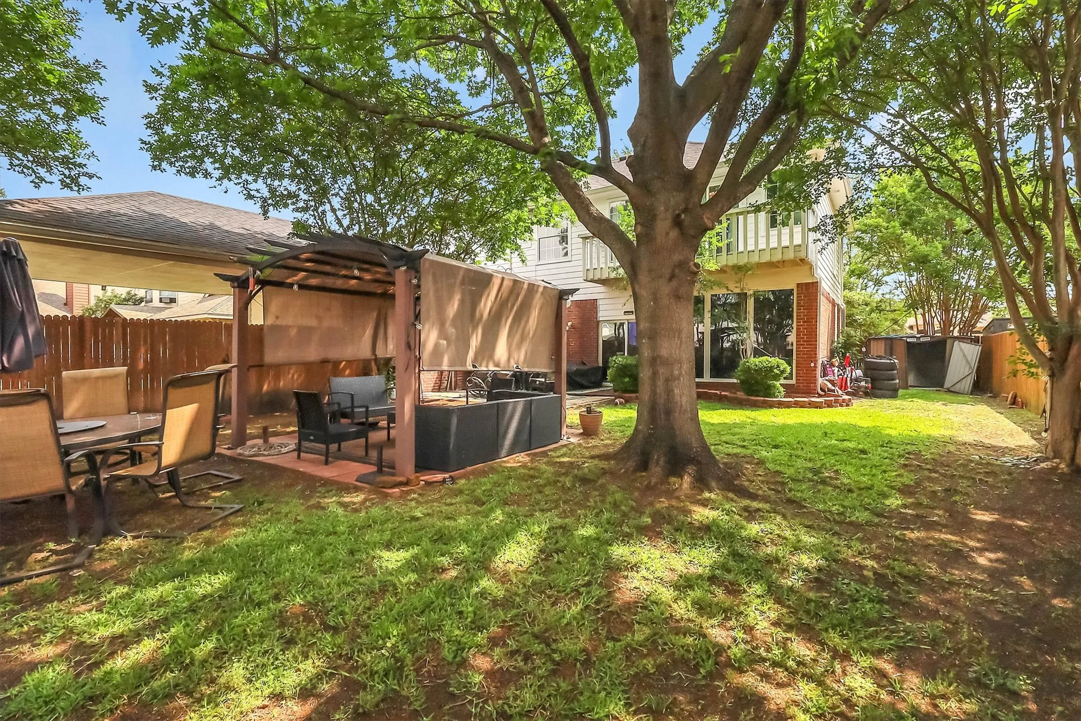 Expansive backyard featuring a large tree, a covered pergola with privacy screens, and a brick patio area