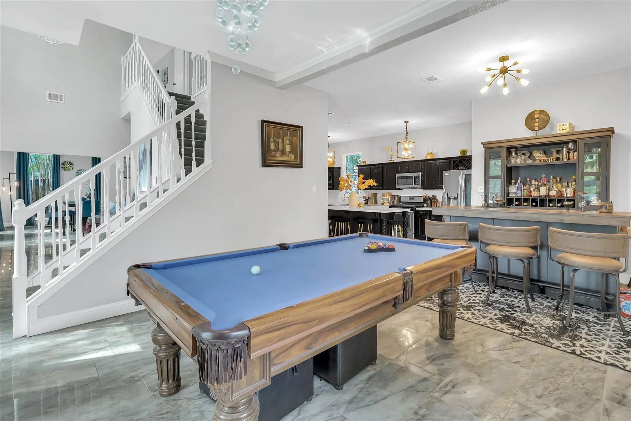 Spacious open-concept interior featuring a billiards area, built-in bar with seating, a prominent staircase with white railings, and large format marble-finish flooring