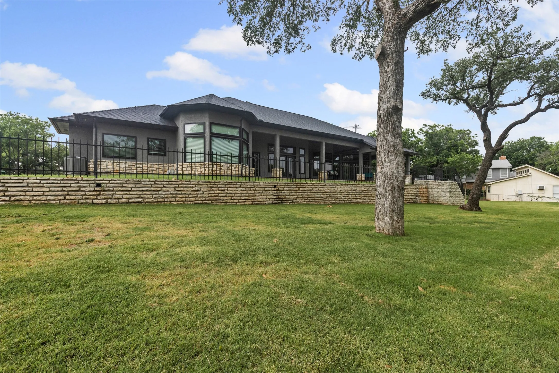 Contemporary stucco exterior with a dark gray roof, multiple large windows, a stone retaining wall, and a covered patio area
