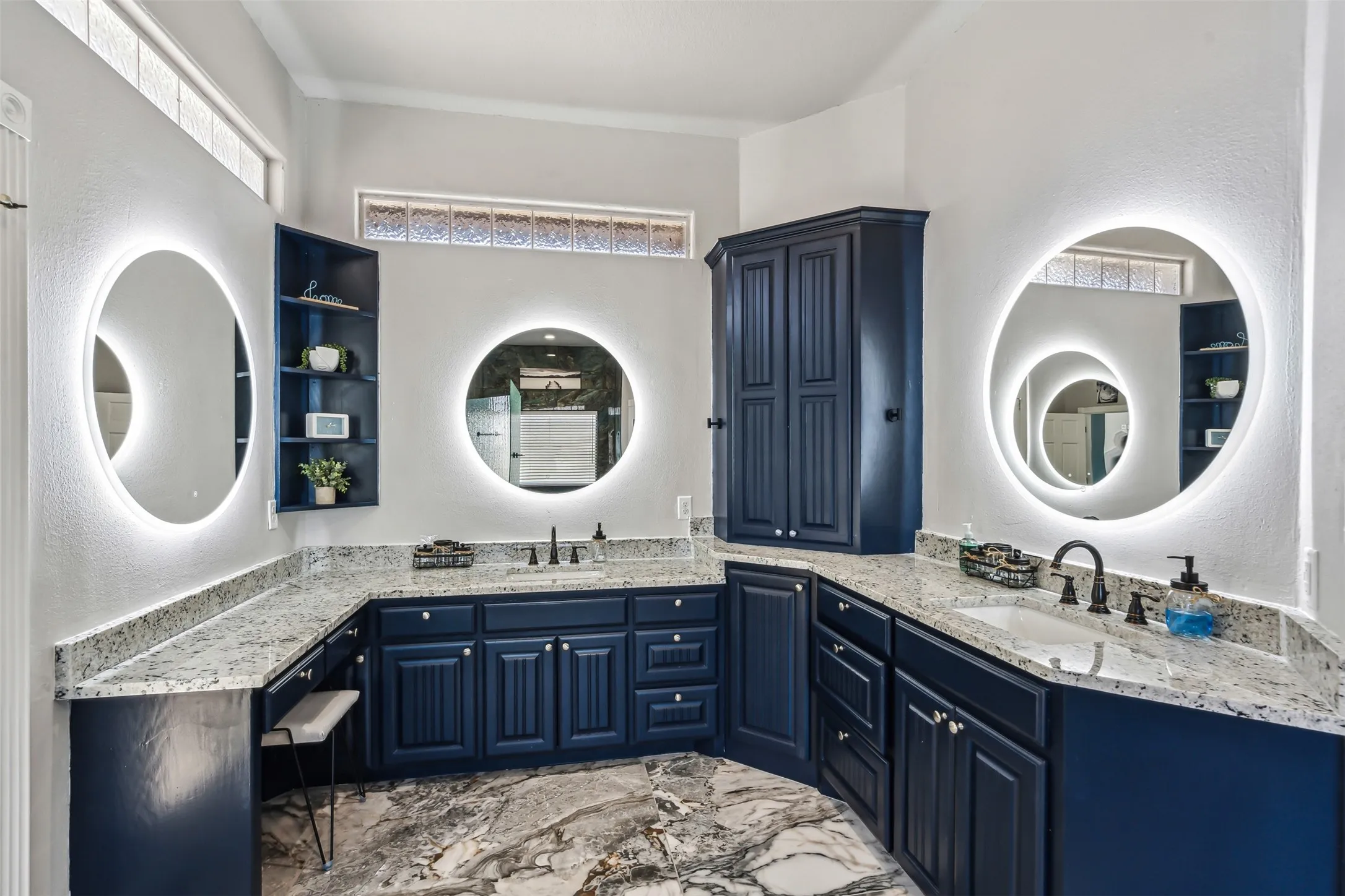 Dual backlit circular mirrors highlight the expansive vanity featuring dark blue cabinetry, light granite-finish countertops, and two undermount sinks