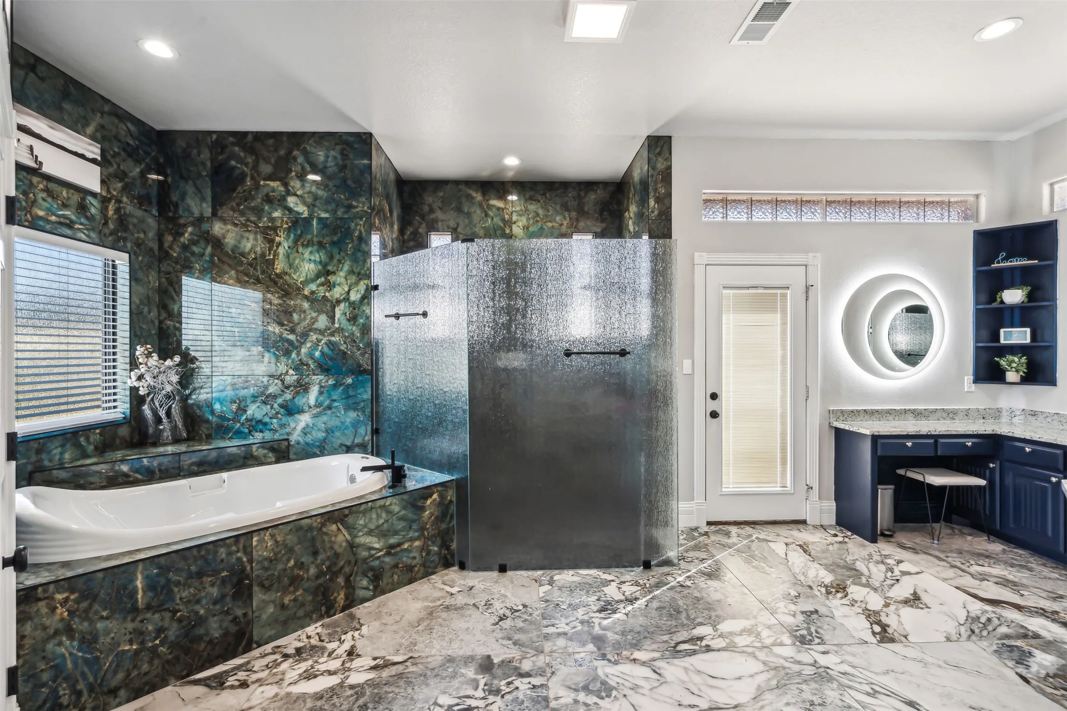 Spacious bathroom featuring patterned wall tile, an elongated soaking tub, a textured glass shower enclosure, a vanity with a dedicated seating area, and a large illuminated mirror
