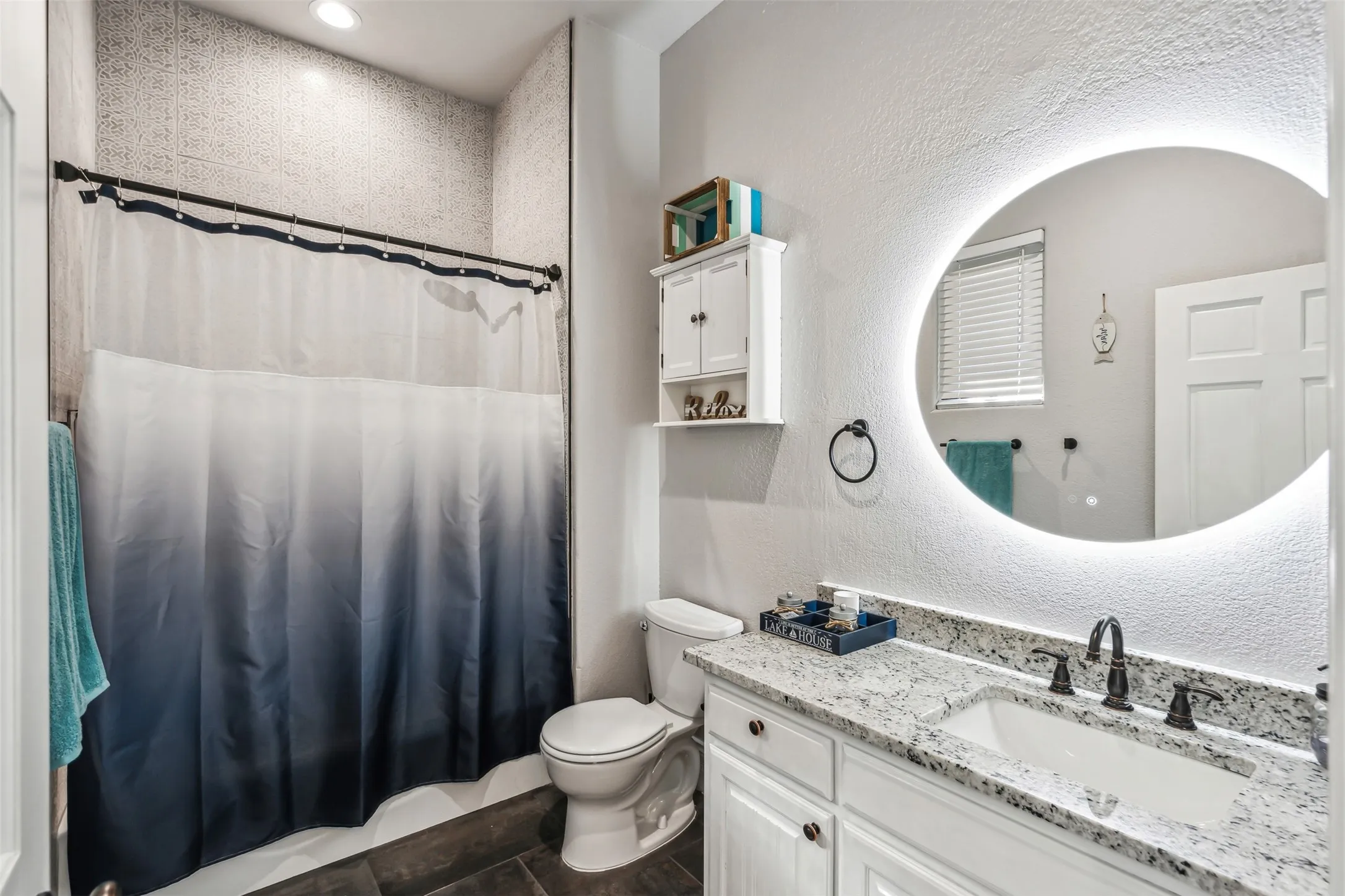 Bathroom featuring a built-in shower with tiled walls, a single vanity with a light-colored countertop, white cabinetry, a built-in toilet, and an illuminated circular mirror