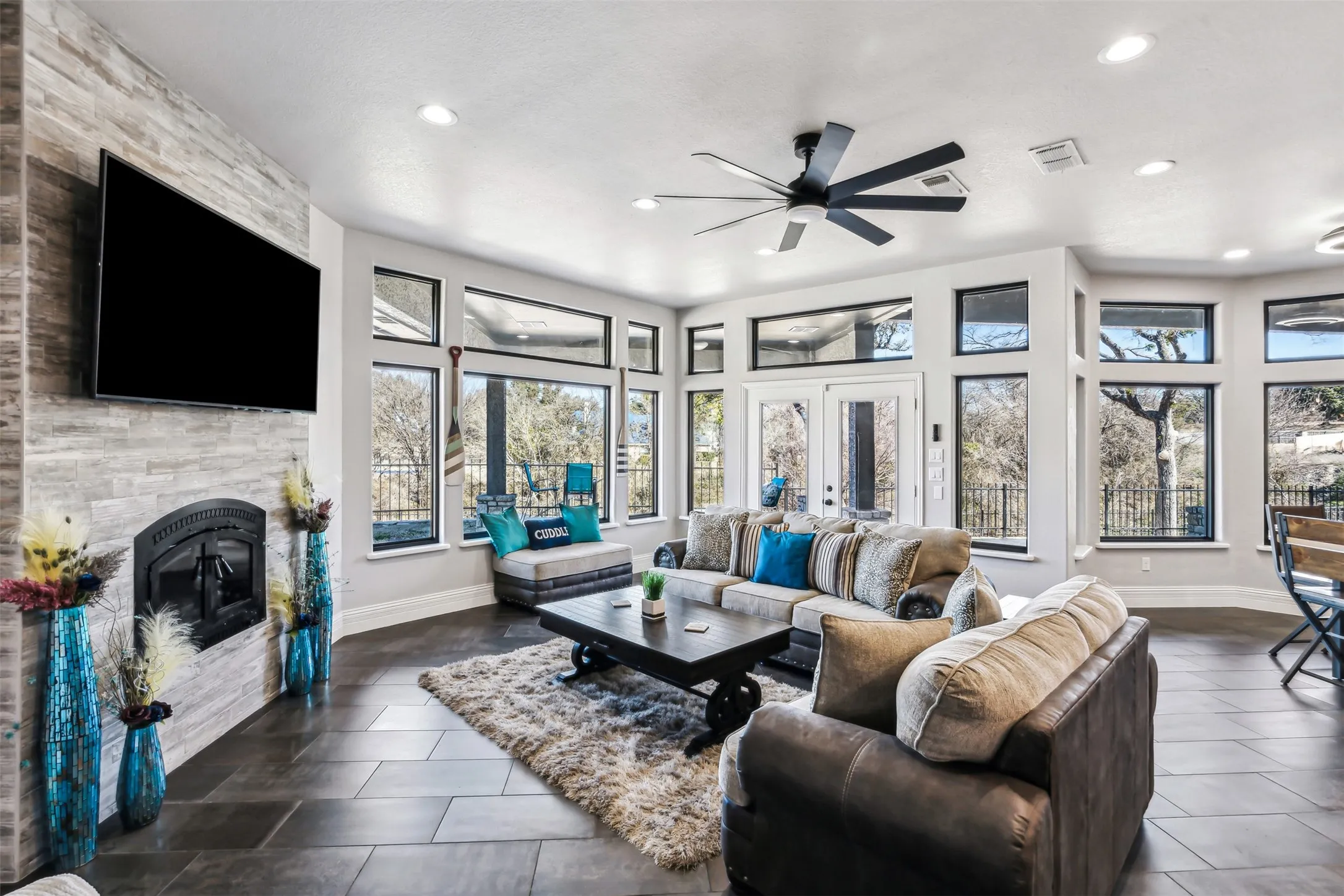 Spacious living area featuring a stacked stone fireplace with an arched firebox, dark tile flooring, a multi-pane glass door opening to an exterior area, and extensive picture windows