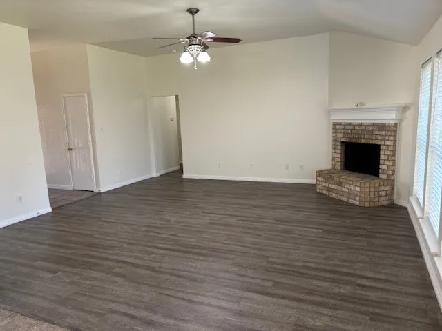 Spacious living area featuring vaulted ceilings, wood-finish flooring, and a corner brick fireplace with a white mantel