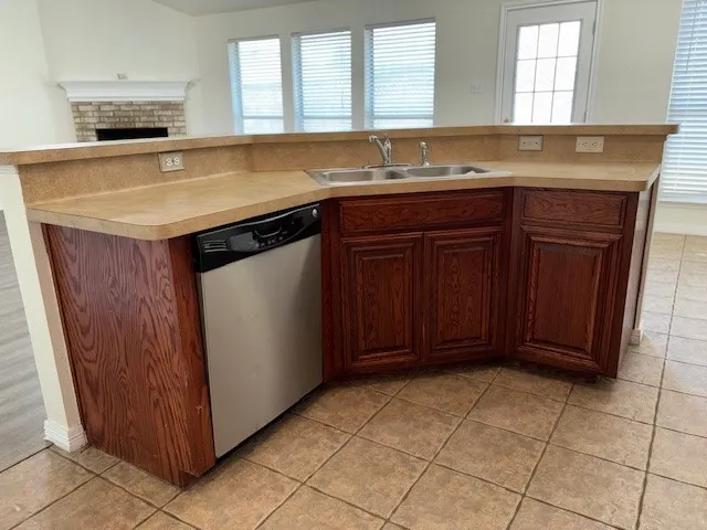Kitchen island with a double basin stainless steel sink, a stainless steel dishwasher, wood cabinetry, and a light-toned countertop
