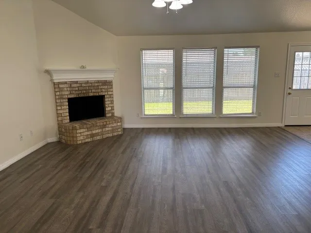 Spacious living area featuring wood-finish flooring, a corner brick fireplace with a white mantle, and three large windows with horizontal blinds