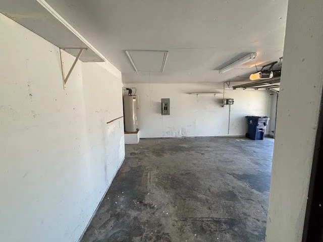 Garage interior featuring a concrete floor, overhead fluorescent lighting, built-in shelving, a water heater, and an electrical panel