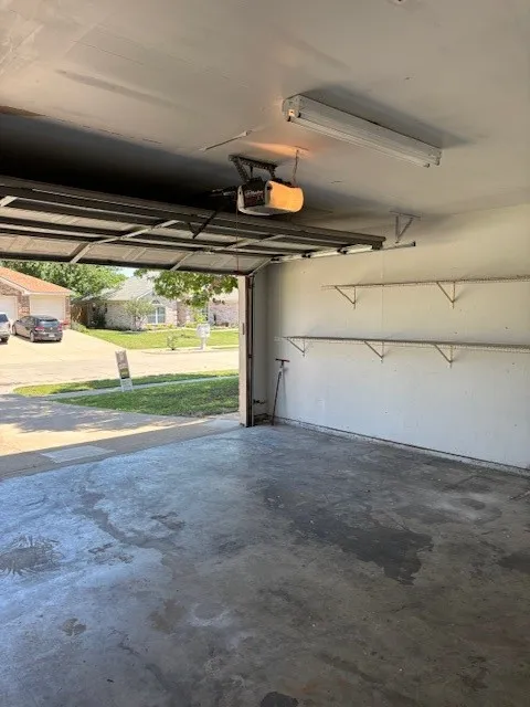 Open garage space featuring a concrete floor, built-in shelving, overhead fluorescent lighting, and an automatic garage door opener