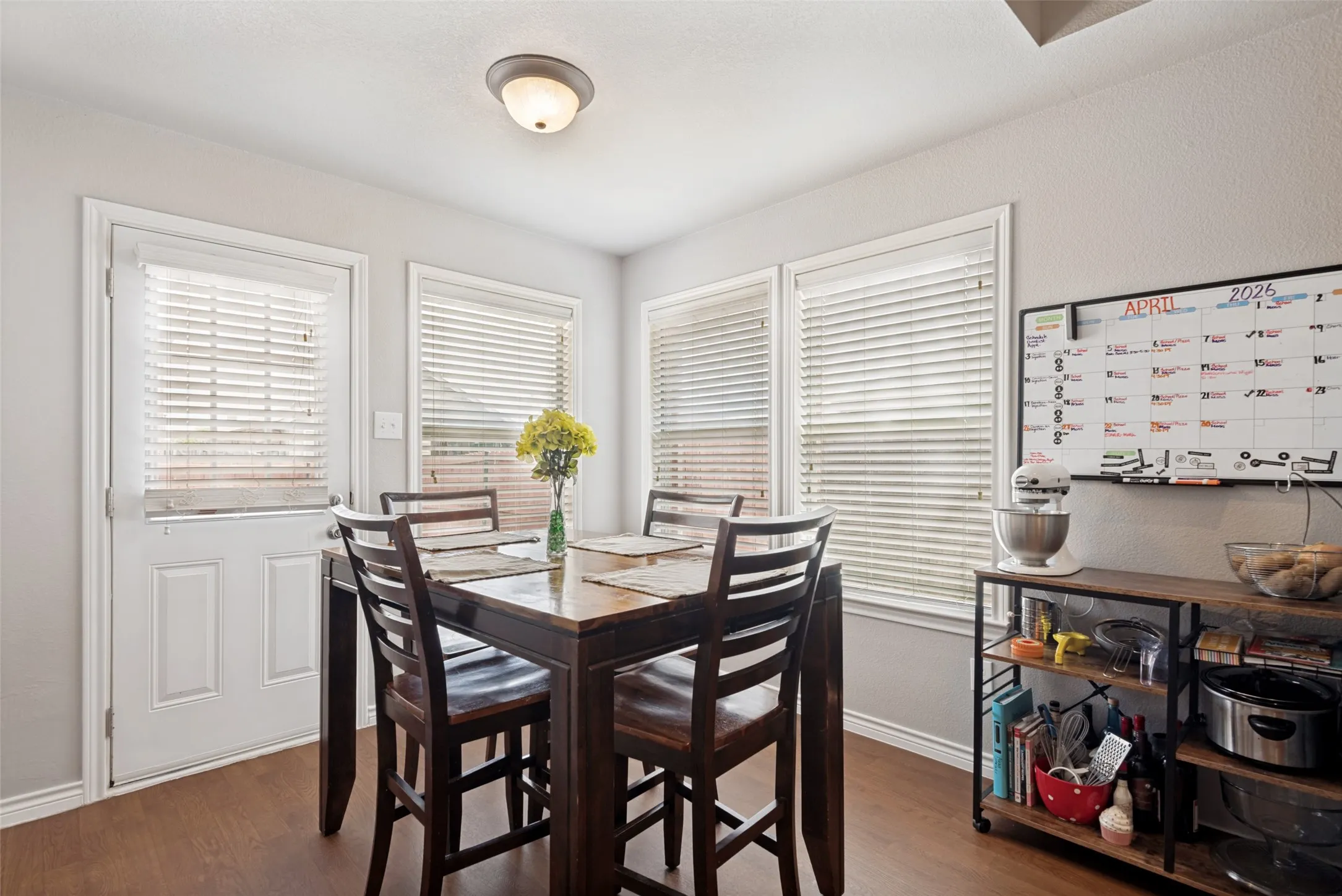 Dining area with warm wood-finish flooring, multiple windows with blinds, a side door, and a flush-mount ceiling light fixture