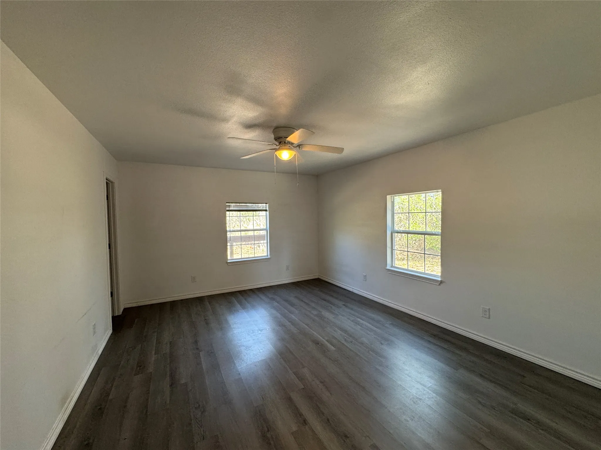 Spacious room featuring wood-finish flooring, white walls, two window openings, a ceiling fan with integrated lighting, and white baseboards