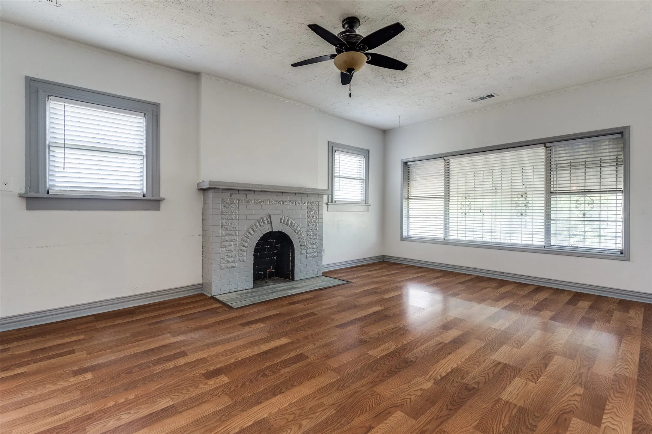 Living area featuring wood-finish flooring, a painted brick fireplace, a ceiling fan, and multiple windows with blinds