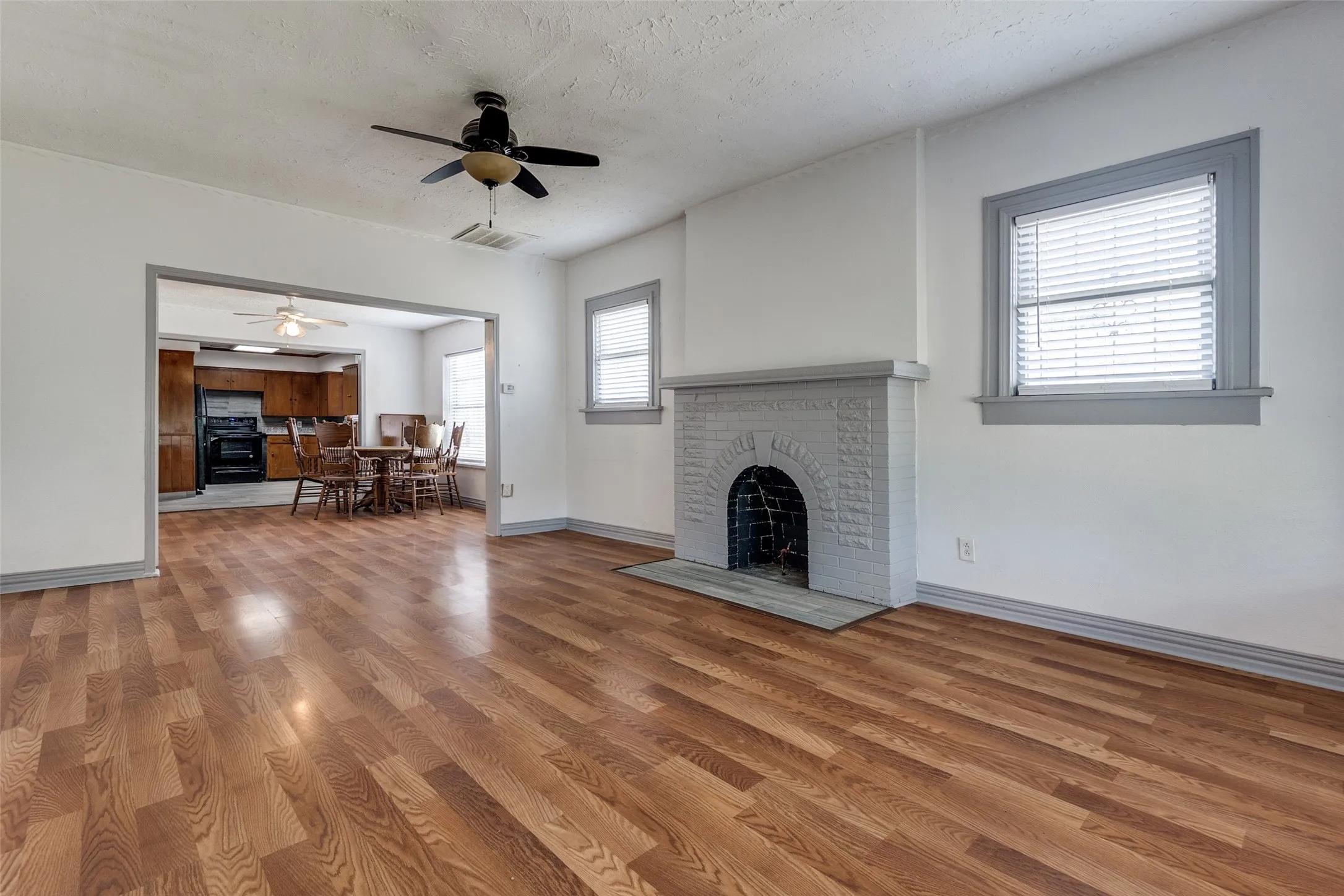 Spacious living area featuring wood-finish flooring, a painted brick fireplace, a ceiling fan, and white walls with contrasting trim