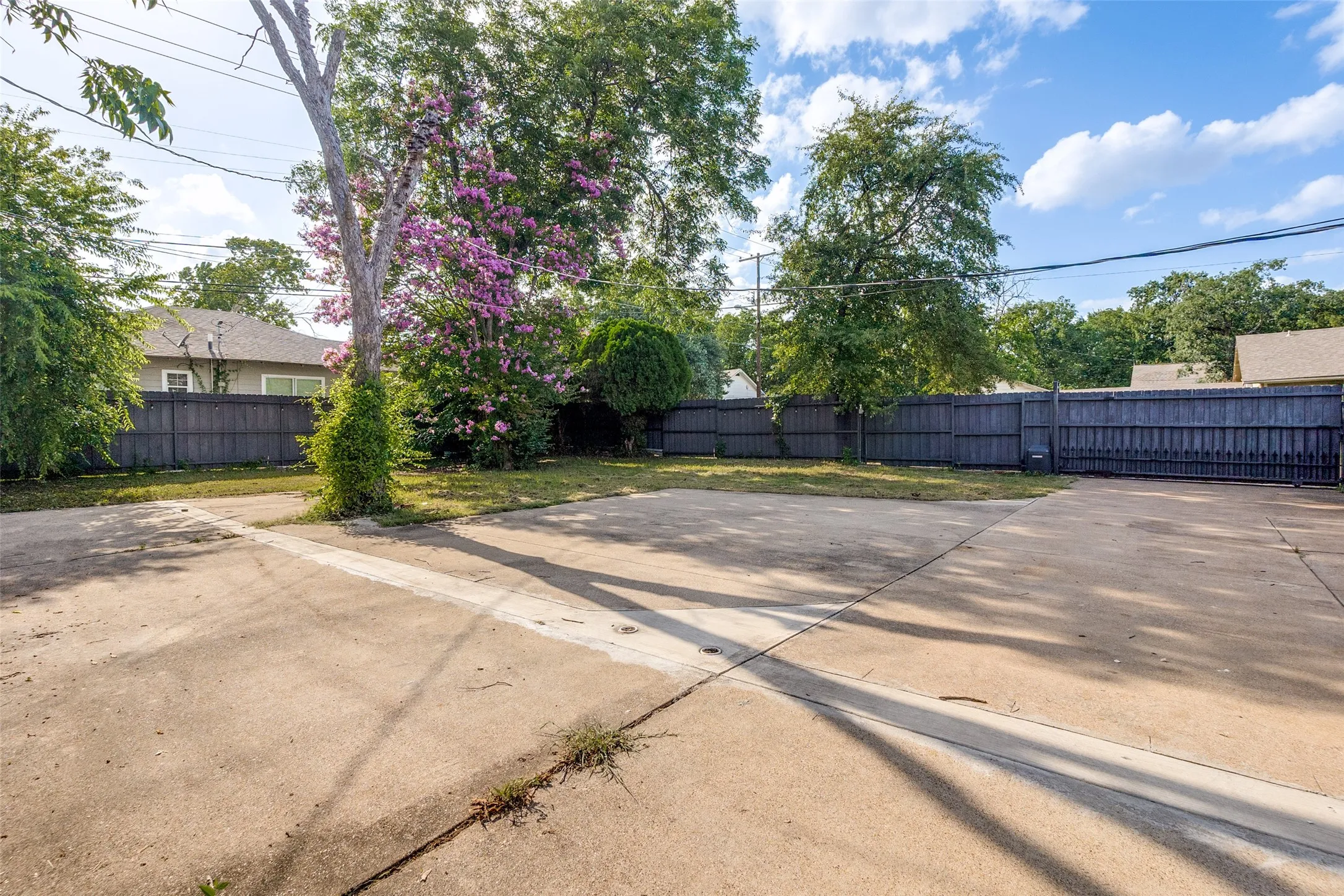 Paved driveway offering ample parking, secured by a tall privacy fence, with mature trees and vibrant flowering bushes providing natural screening