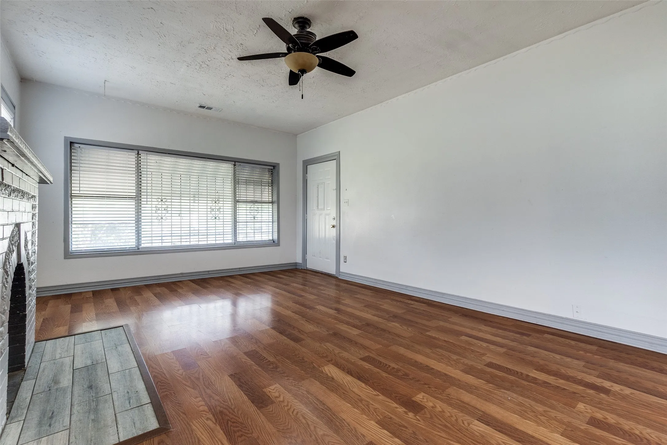 Expansive living area with wood-finish flooring, a decorative fireplace with a tile hearth, and a multi-pane window with blinds