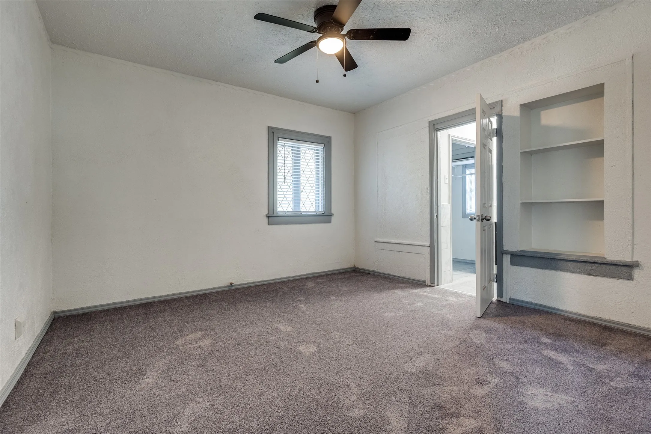 Spacious room featuring neutral carpeting, a ceiling fan with integrated lighting, a window with horizontal blinds, built-in shelving, and a white door with a silver-toned knob