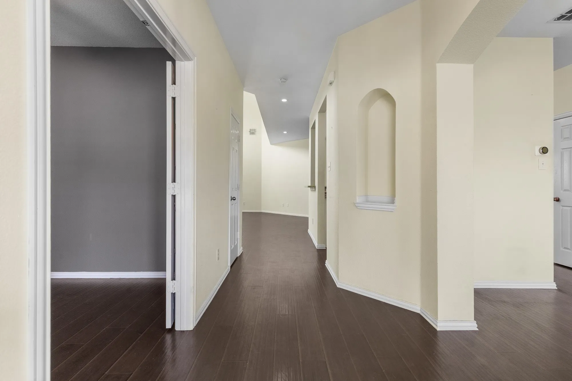 Expansive hallway featuring wood-finish flooring, light-toned wall paint, white baseboards, a decorative wall niche, and an arched doorway