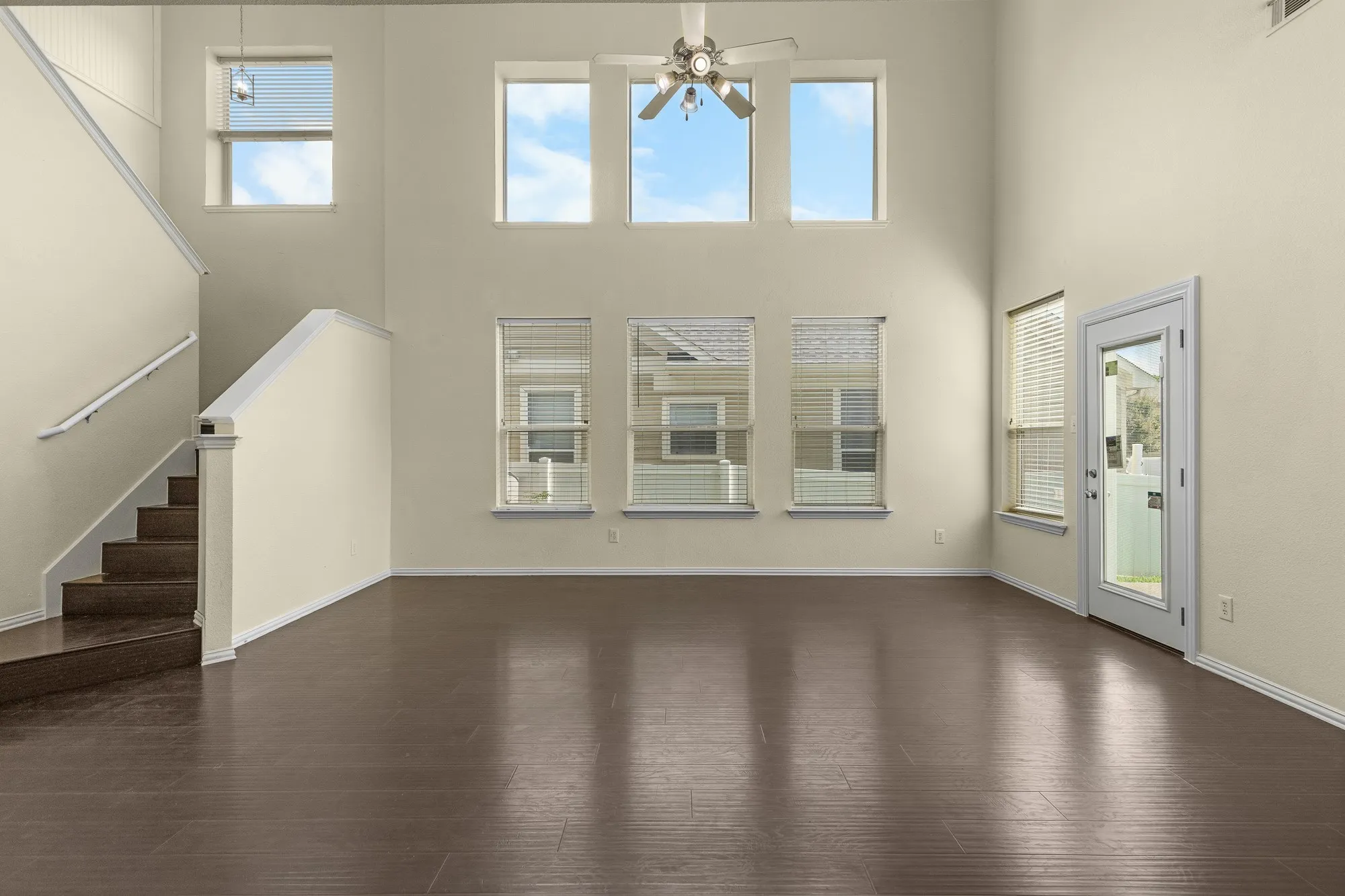 Spacious living area featuring high ceilings, wood-finish flooring, a ceiling fan, multiple windows with blinds, and a glass-paneled exterior door