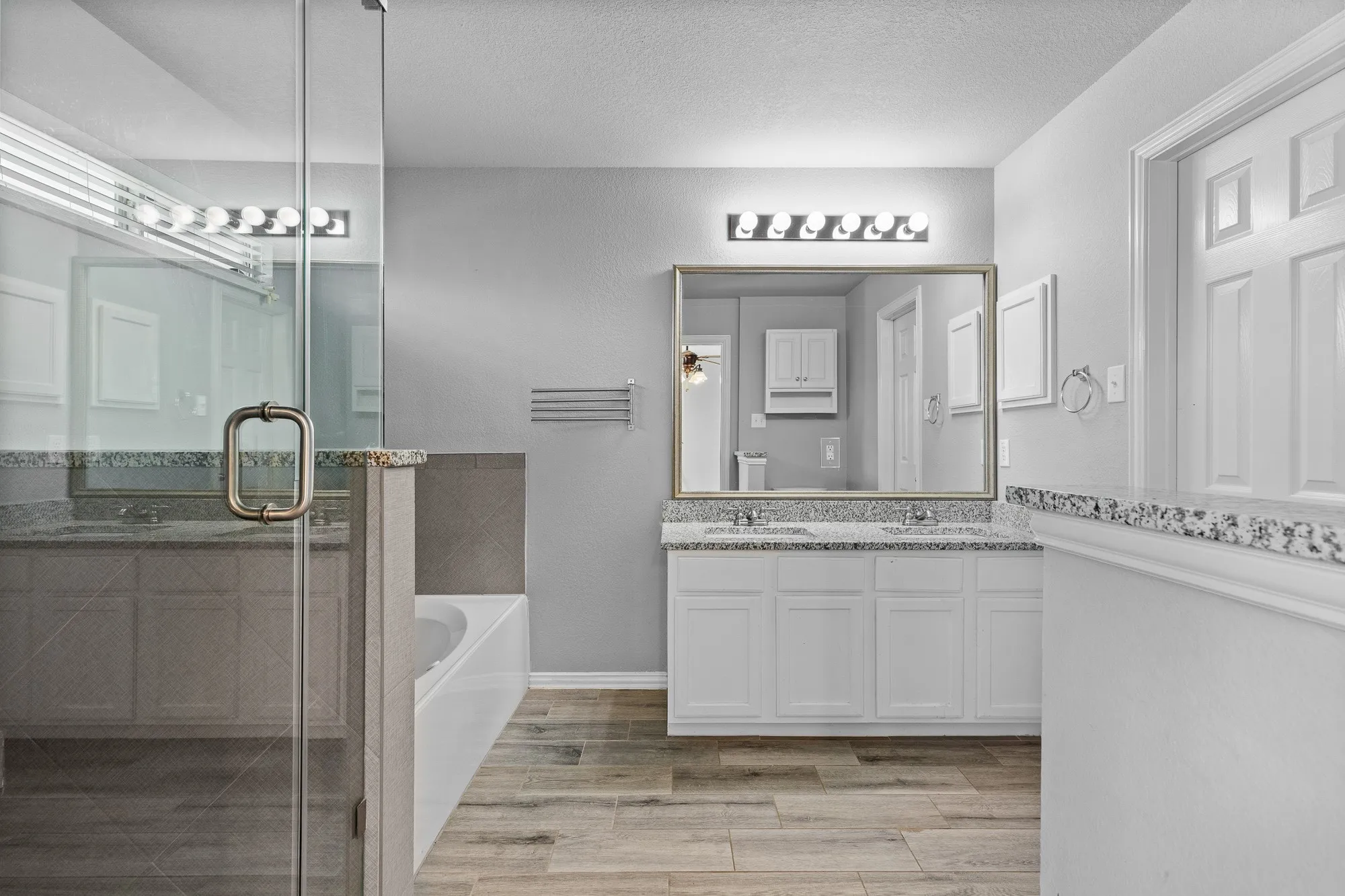 Bathroom featuring a dual vanity with granite countertops, wood-finish flooring, a frameless glass shower enclosure, and a separate soaking tub