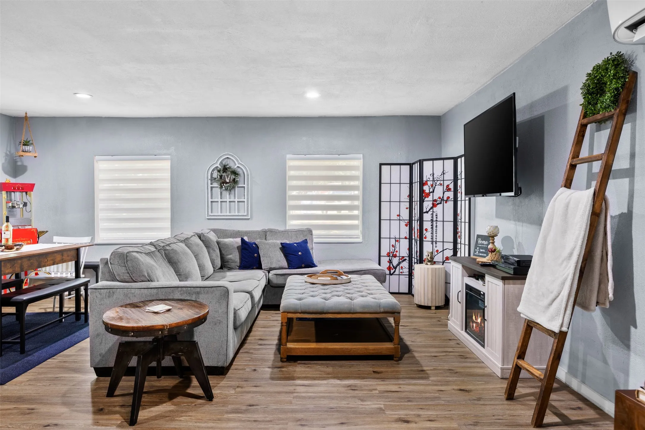 Open-concept interior featuring wood-finish flooring, two windows with horizontal blinds, a wall-mounted television, and an entertainment console with an electric fireplace