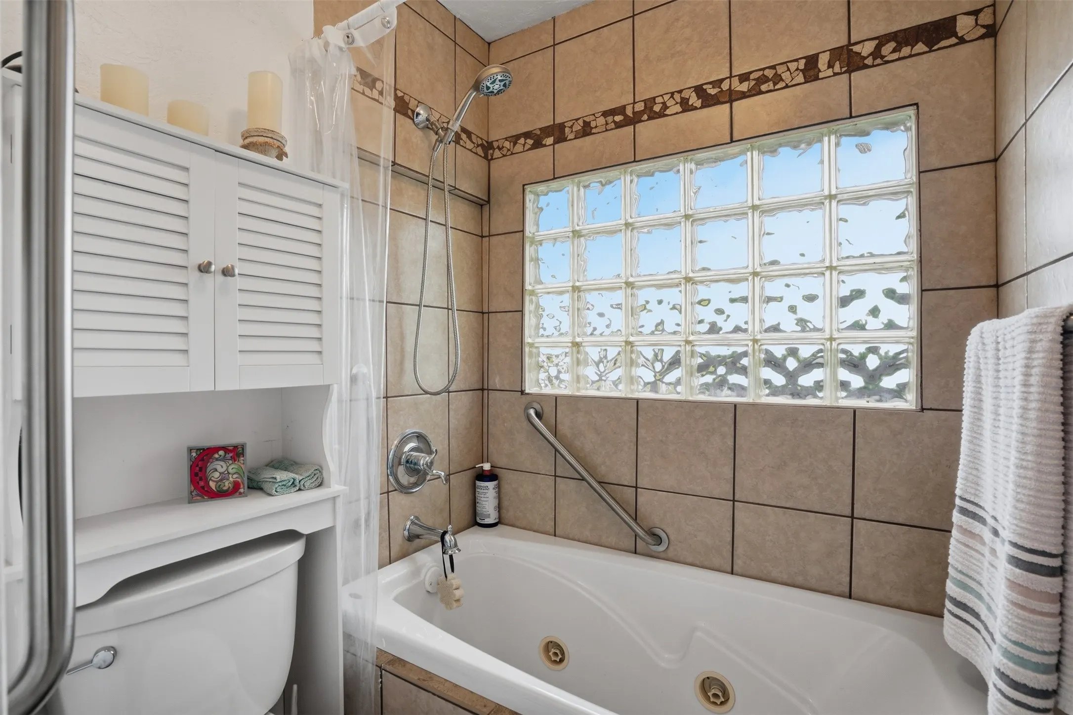 Tiled bathtub surround featuring a glass block window, a shower head with handheld sprayer, an integrated grab bar, and a white over-the-toilet cabinet with louvered doors