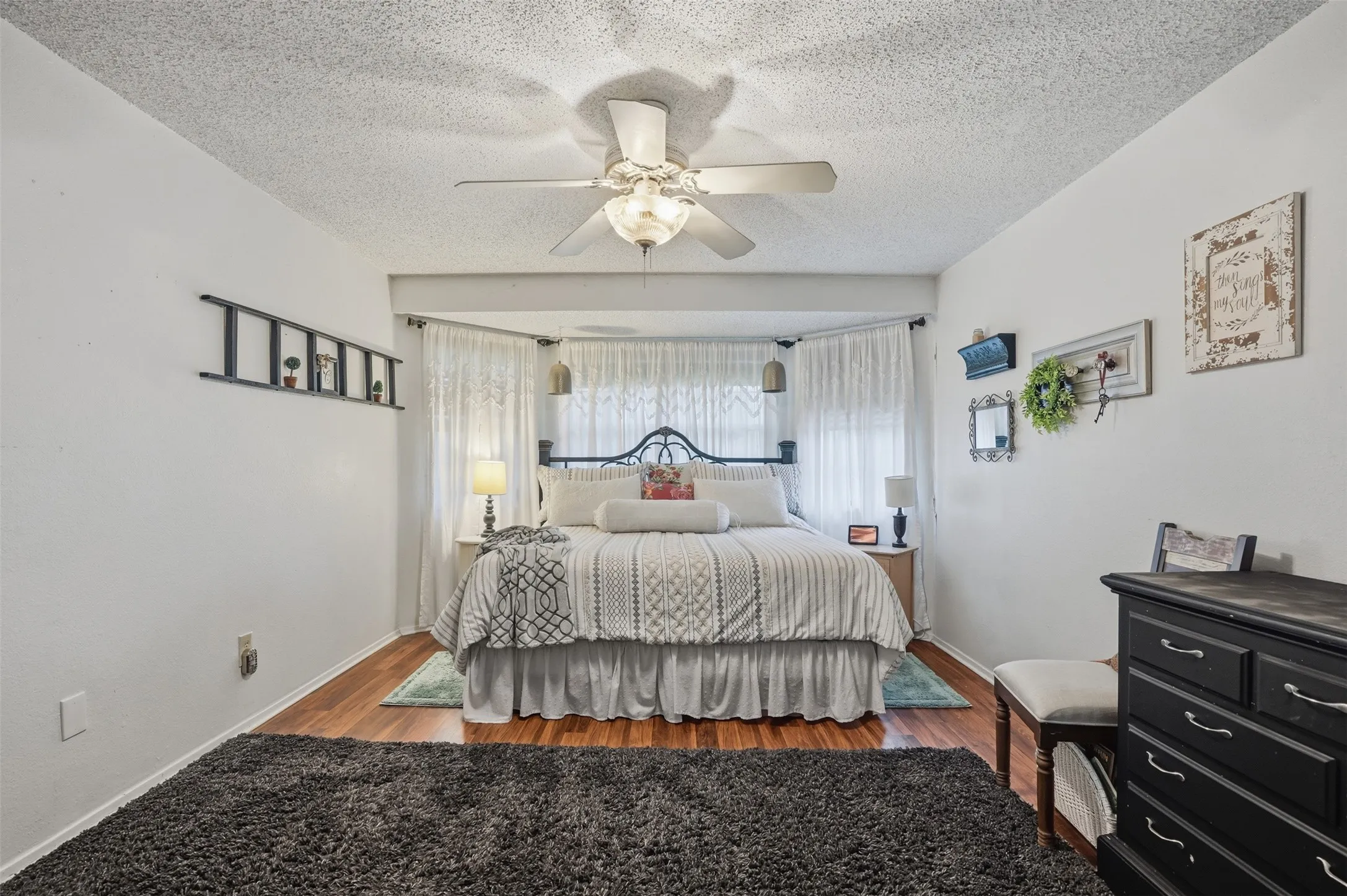 Recessed bedroom area featuring wood-finish flooring, a ceiling fan with integrated lighting, and a window dressed with sheer white curtains