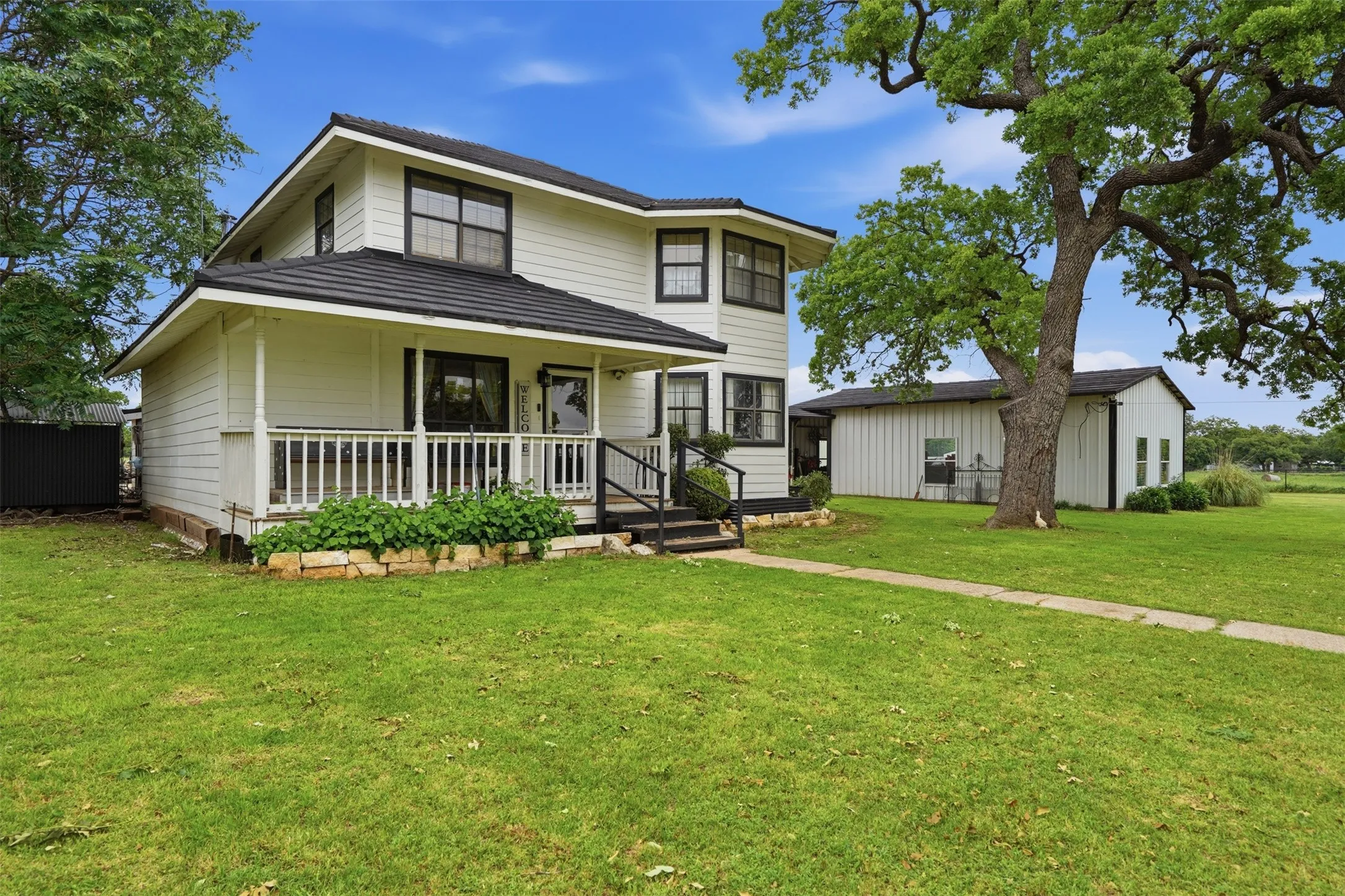 Two-story residence featuring white siding and black trim, a covered porch with white railings, a stone-lined garden bed, a paved walkway, and a separate outbuilding with a black roof