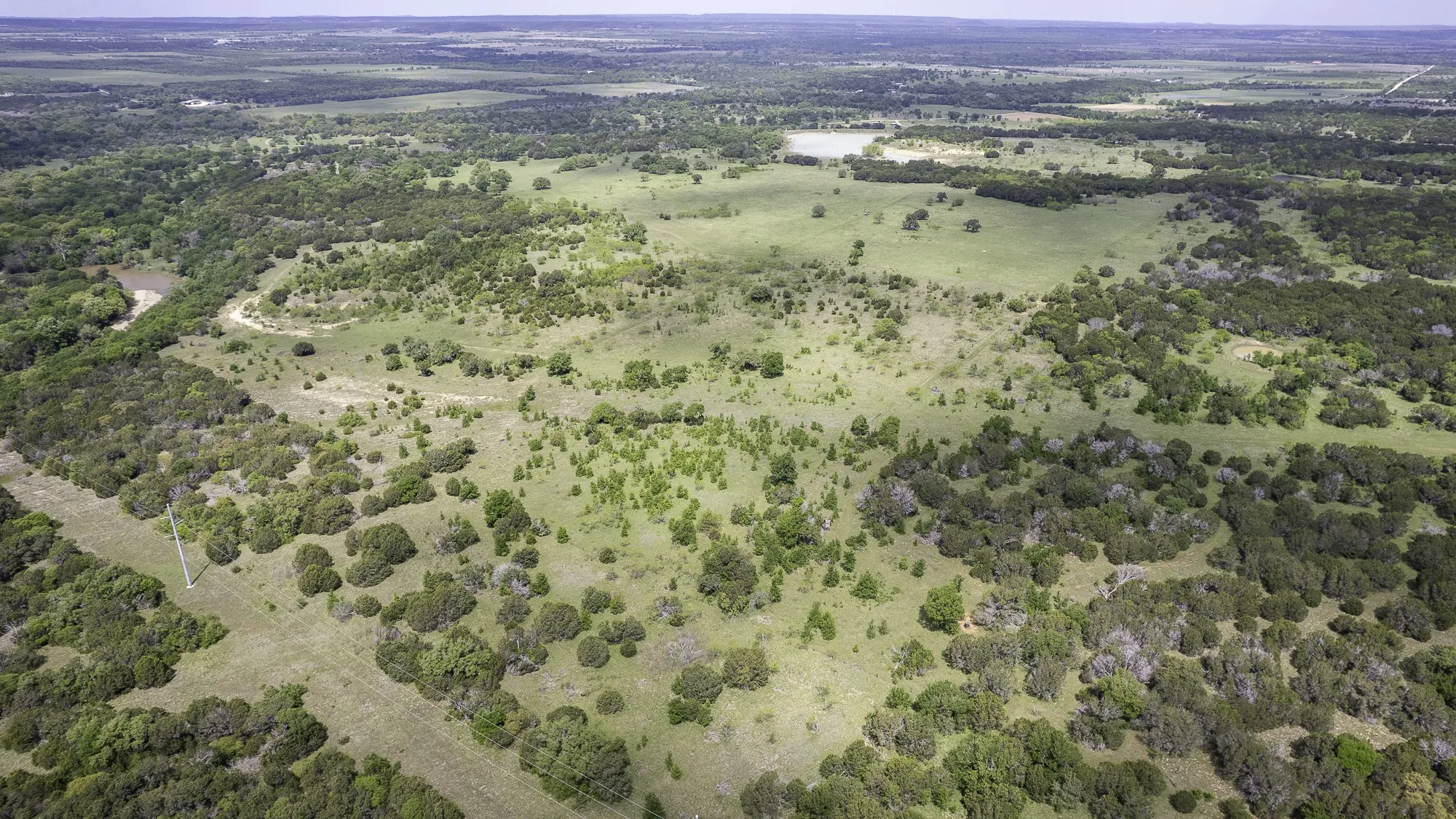 Expansive aerial view showcasing diverse terrain, including areas of dense tree coverage, open grassy fields, and a visible lake