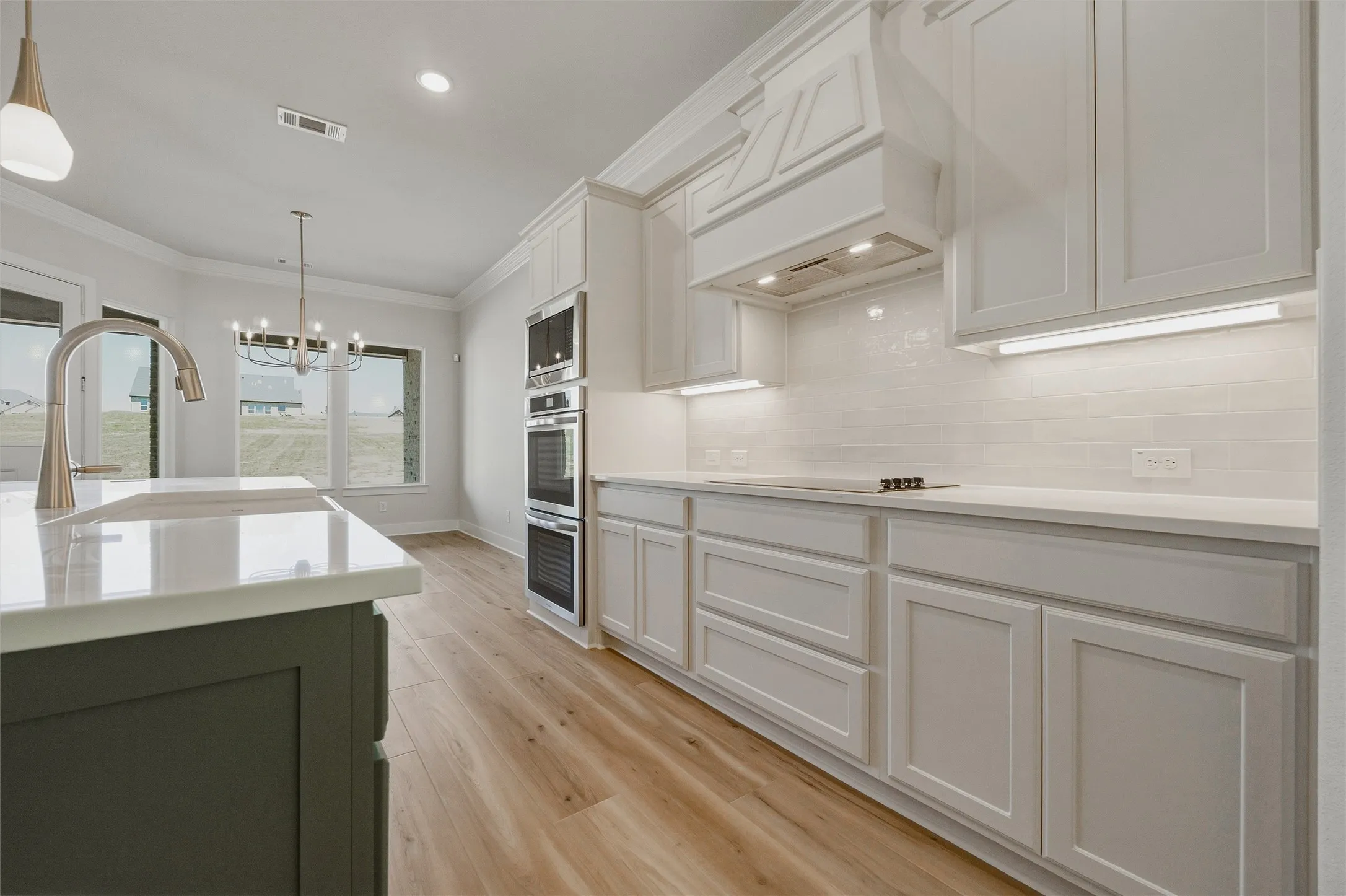 Kitchen featuring white shaker-style cabinetry, double wall ovens, a cooktop, wood-finish flooring, and a large central island with a sink and pendant lighting