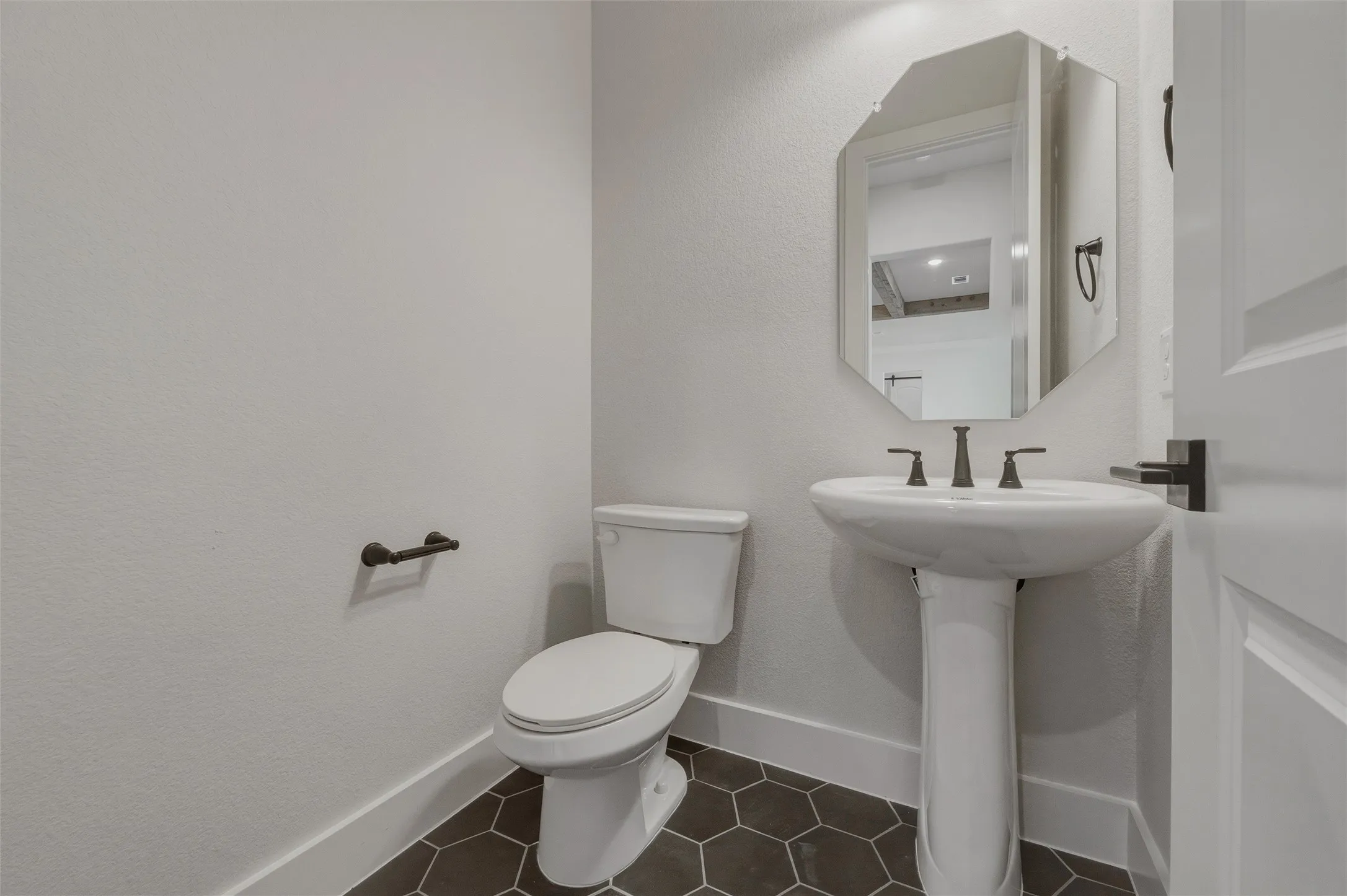 Pedestal sink with dark-finished faucet, octagonal mirror, toilet, and dark hexagon tile flooring
